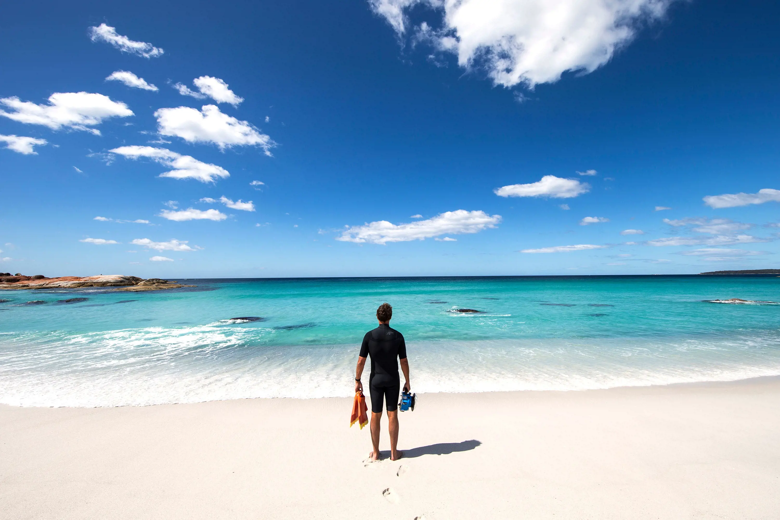 A person in a wetsuit stands on bright white pristine sand, holding flippers and snorkelling goggles. He looks out to the water, which is clear aqua blue.