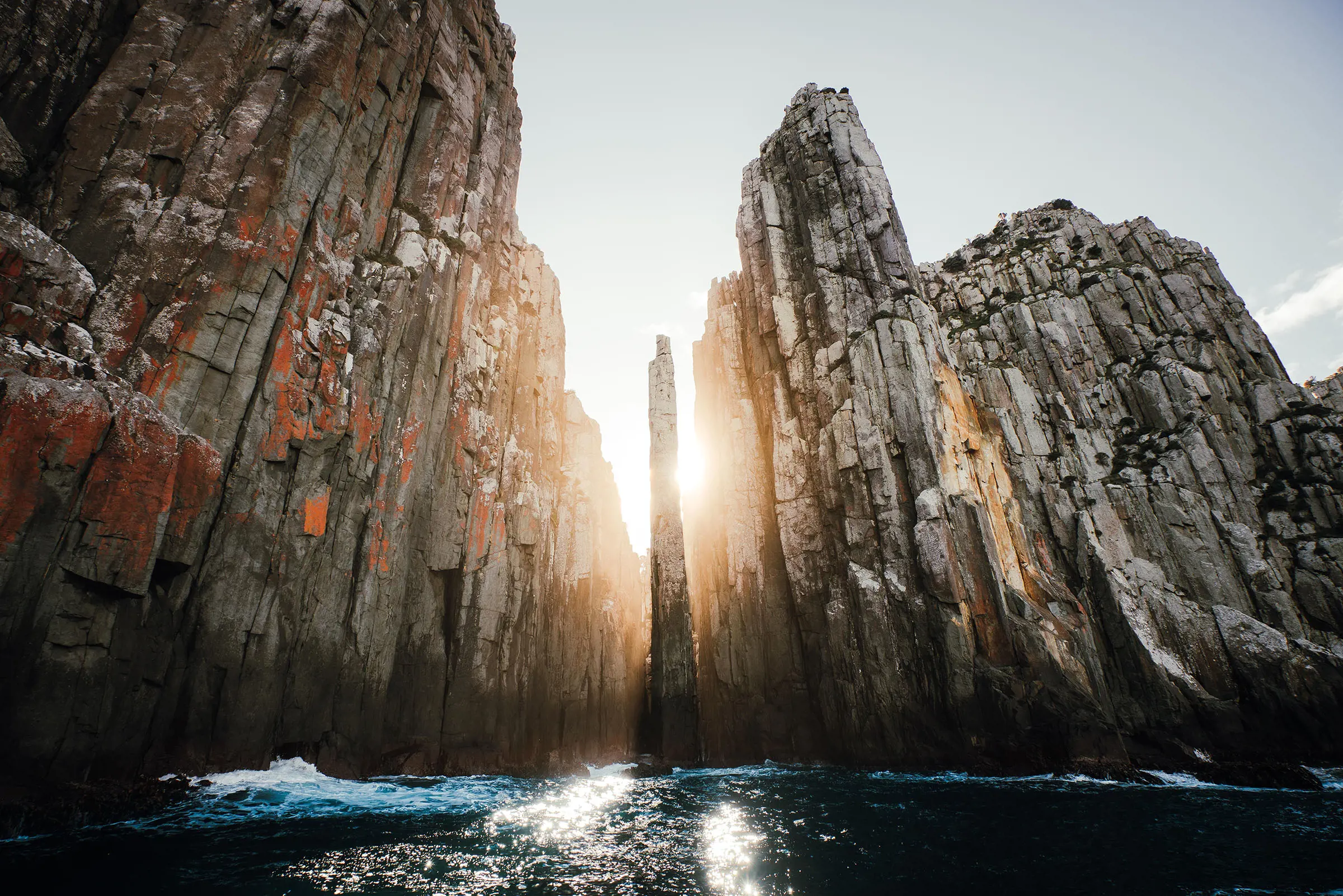 Vertical dolerite sea cliffs rise sharply up from the water, some with an orange hue from growing lichen. The sun shines brightly between two cliffs.