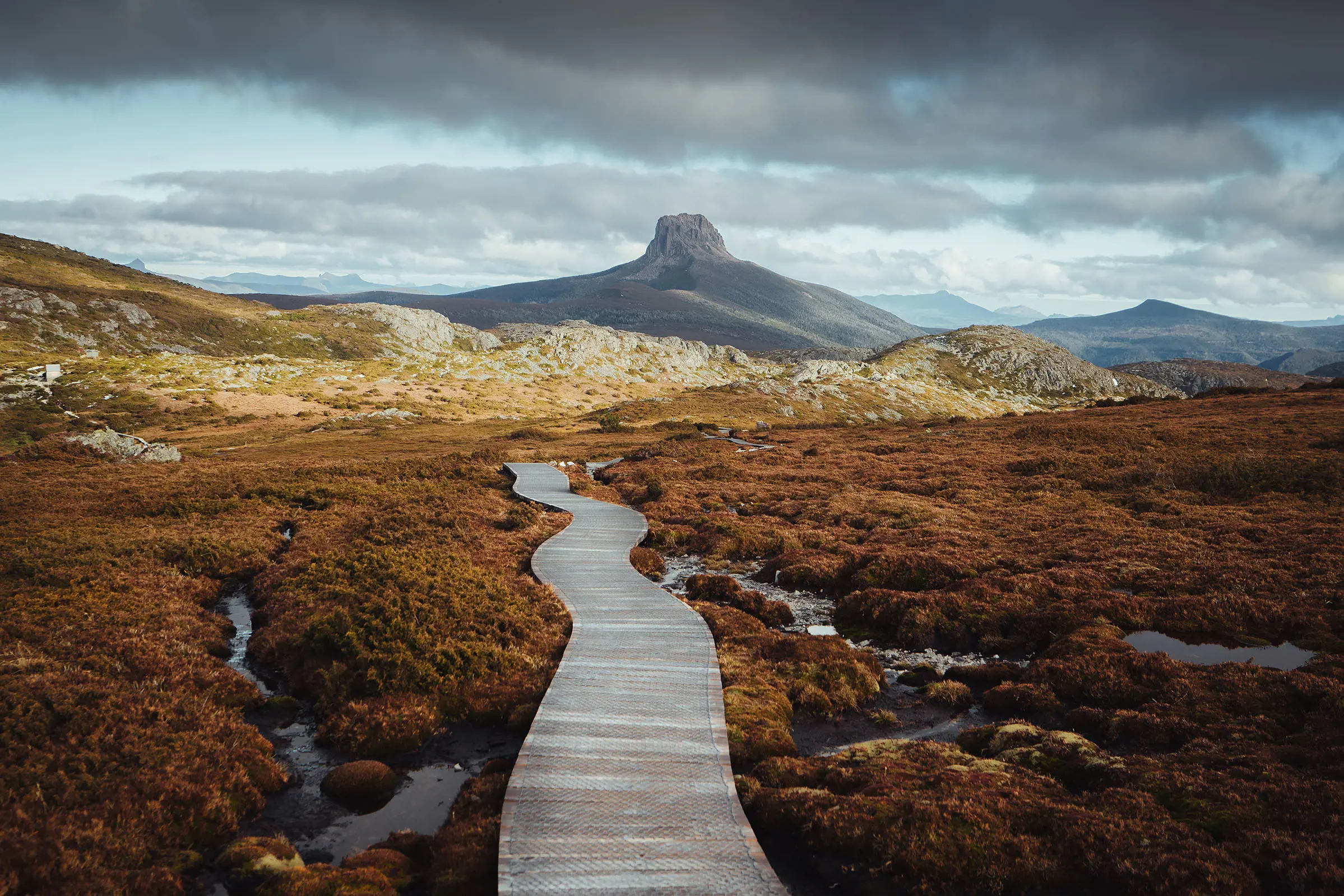 Landscape of the Overland Track, with Barn Bluff in the backdrop. The leaves are orange Autumn coloured and a wooden duckboard snakes its way along the ground.