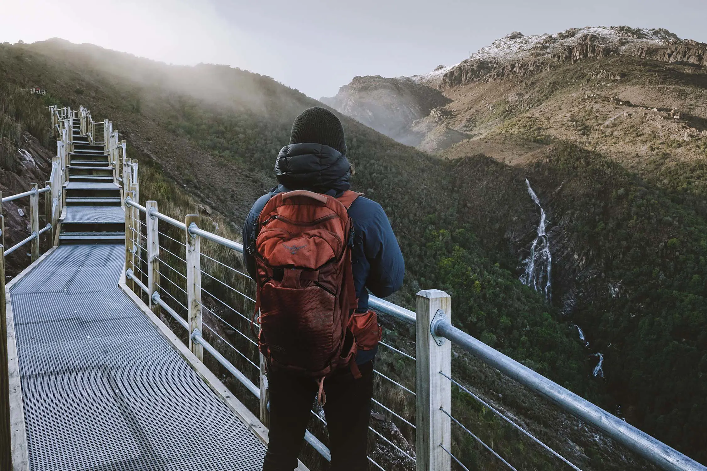 A person with a red backpack stands on a metal walkway, on the side of a barren mountain. In the distance, a very thin waterfall cascades down into the bush below.
