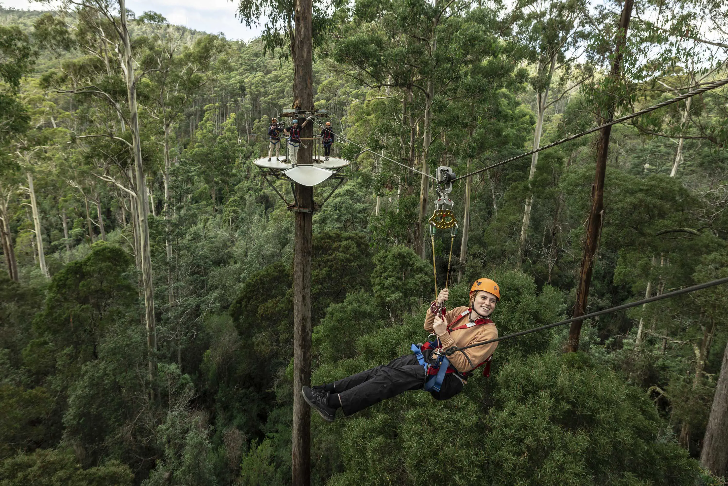 A teenage girl hangs from the harness of a zipline travelling high above green forest canopy.