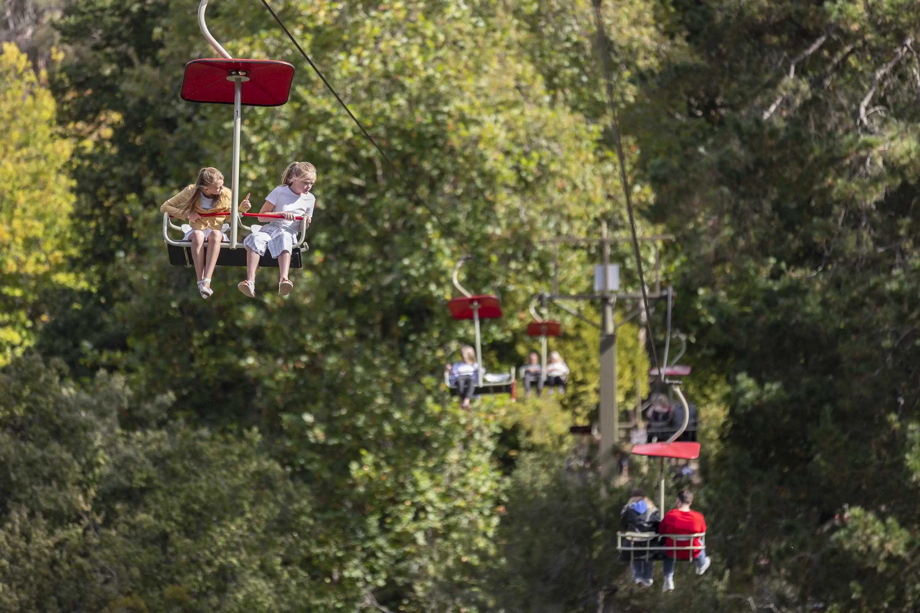 Two young girls sit in the chairlift that traverses a large green field, parklands and water. More chair lifts can be seen travelling in the opposite direction.