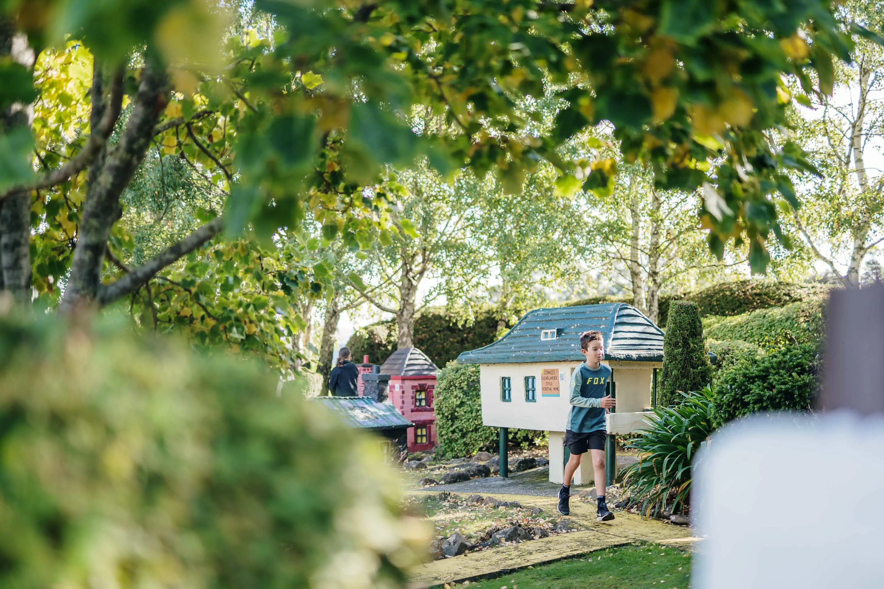 A young boy runs along a path lined with colourful miniature houses under the shade of trees.