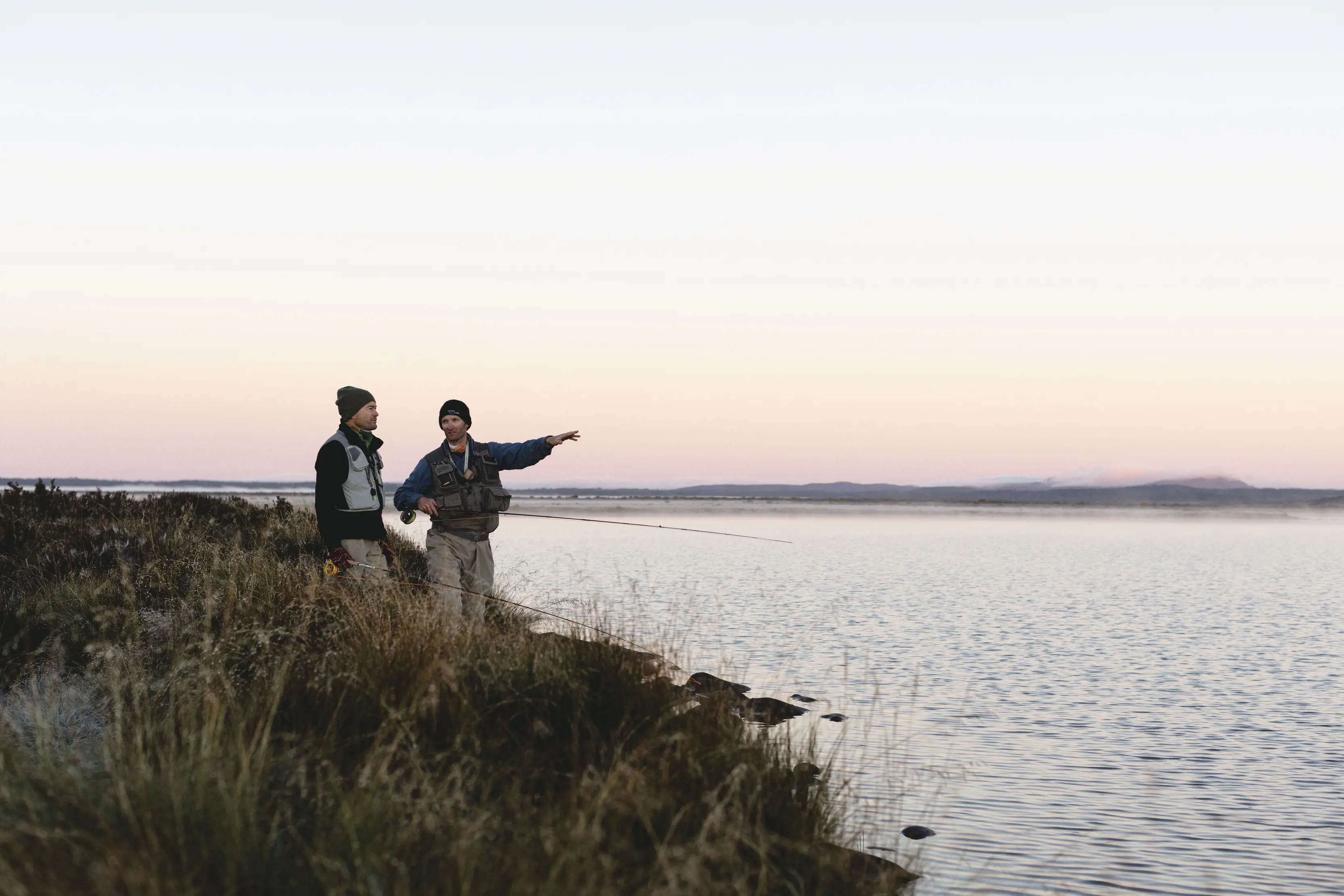 Two men in beanies stand on the flat scrubby bank of a very wide body of water. The sky is the slight pink, orange and yellow fade of sunset.