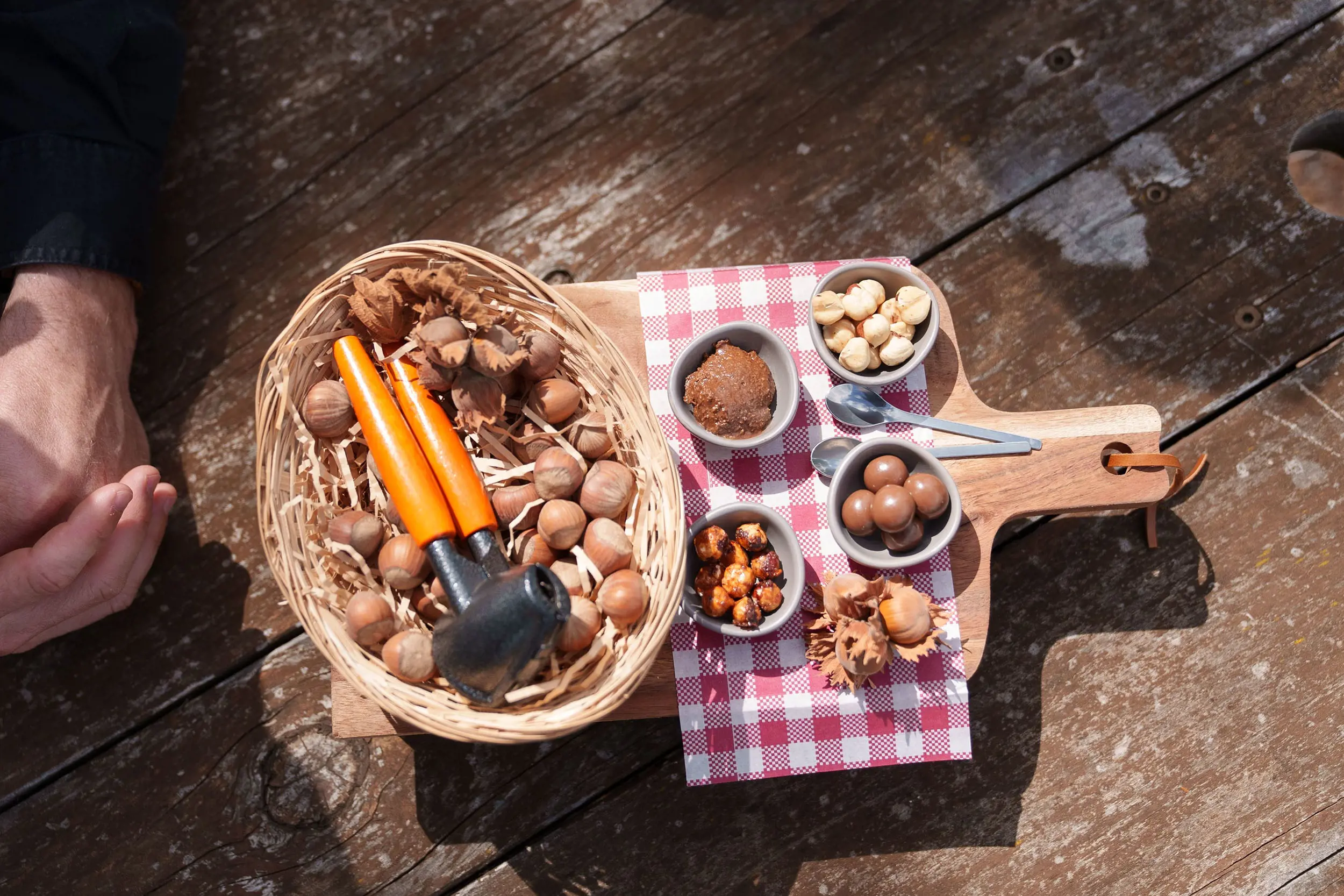 A top-down view of a wooden paddle, carrying a basket of hazelnuts and a nutcracker, and smaller bowls with pieces of cracked hazelnuts.