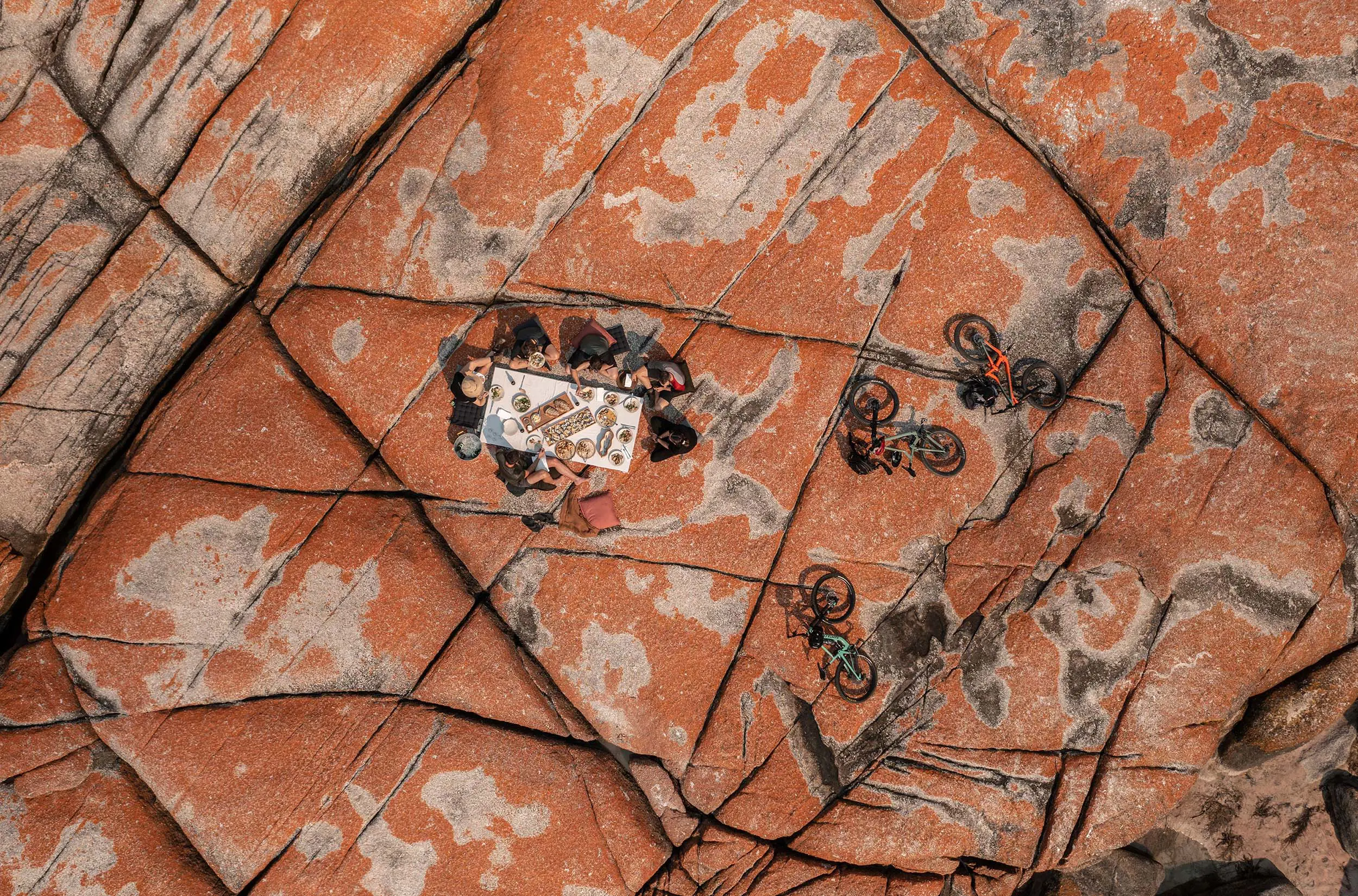 A top-down view of a group of people having a picnic on some vividly orange coloured rocks. Nearby, a few mountain bikes are laid on their sides.