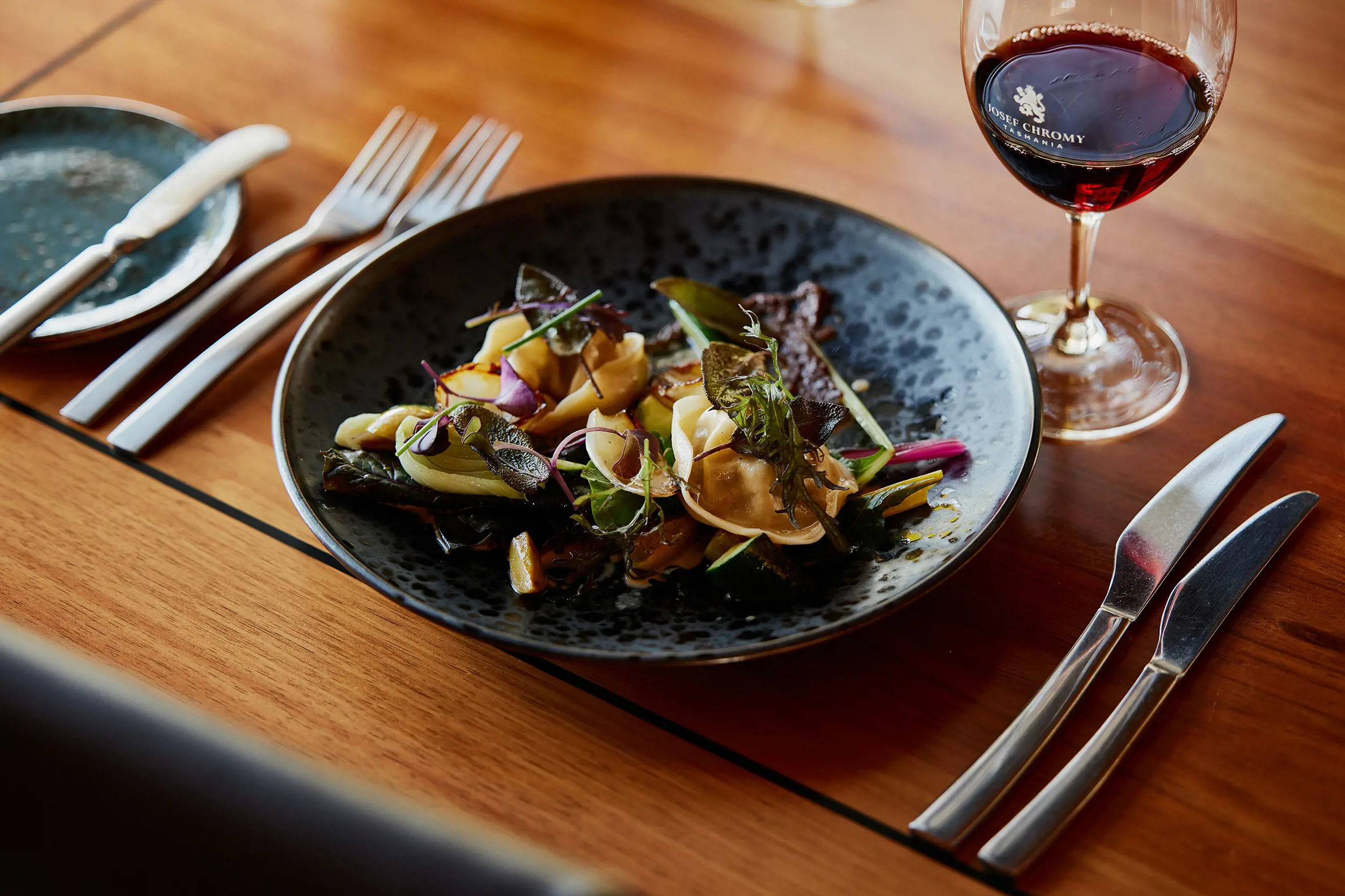 An artfully plated meal sitting on a wooden table. Two sets of cutlery and a glass of red wine are laid out.