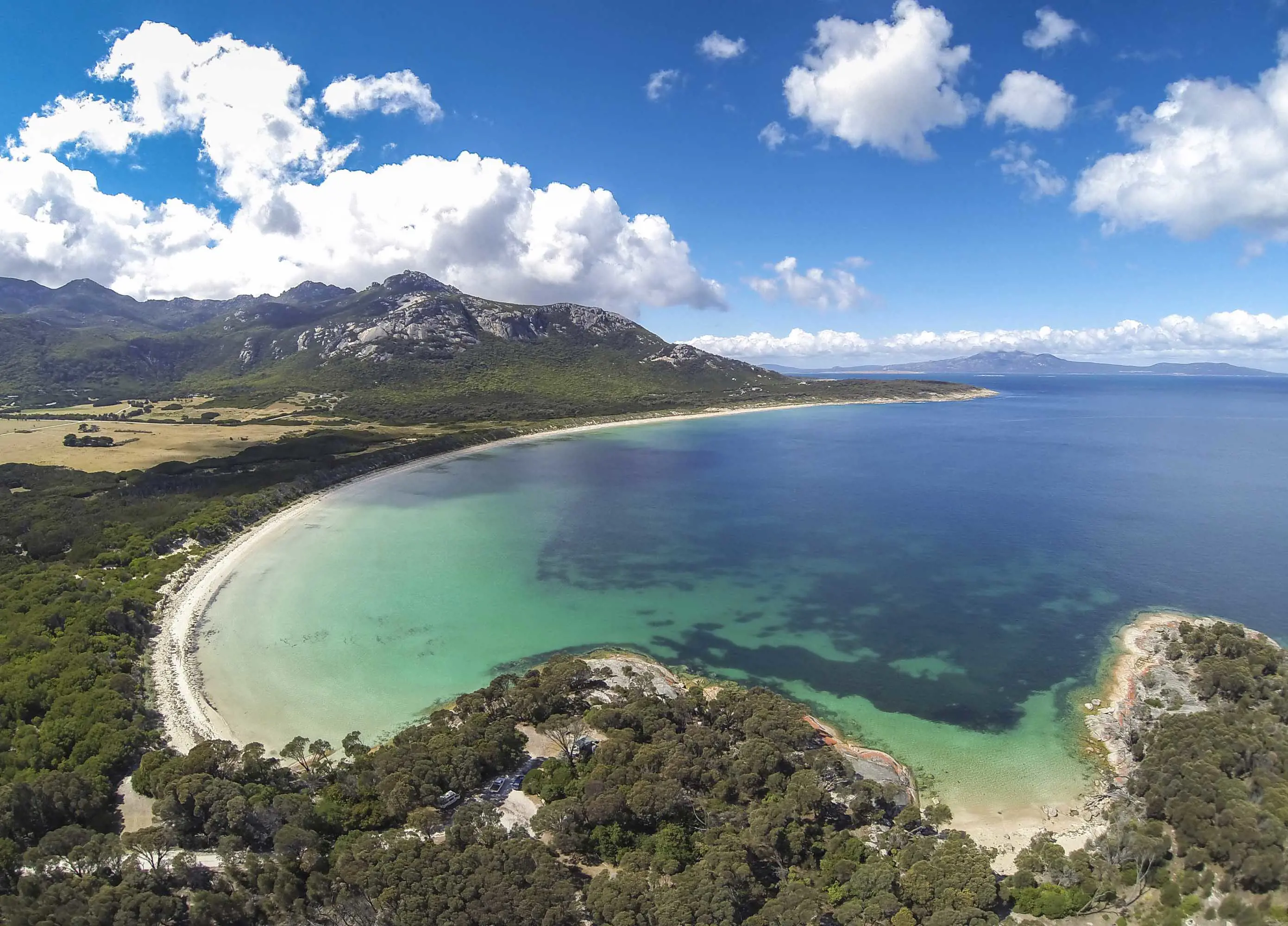 A sweeping bay with rocky outcrops, white sand beaches and turquoise waters on a sunny day with patchy white clouds.