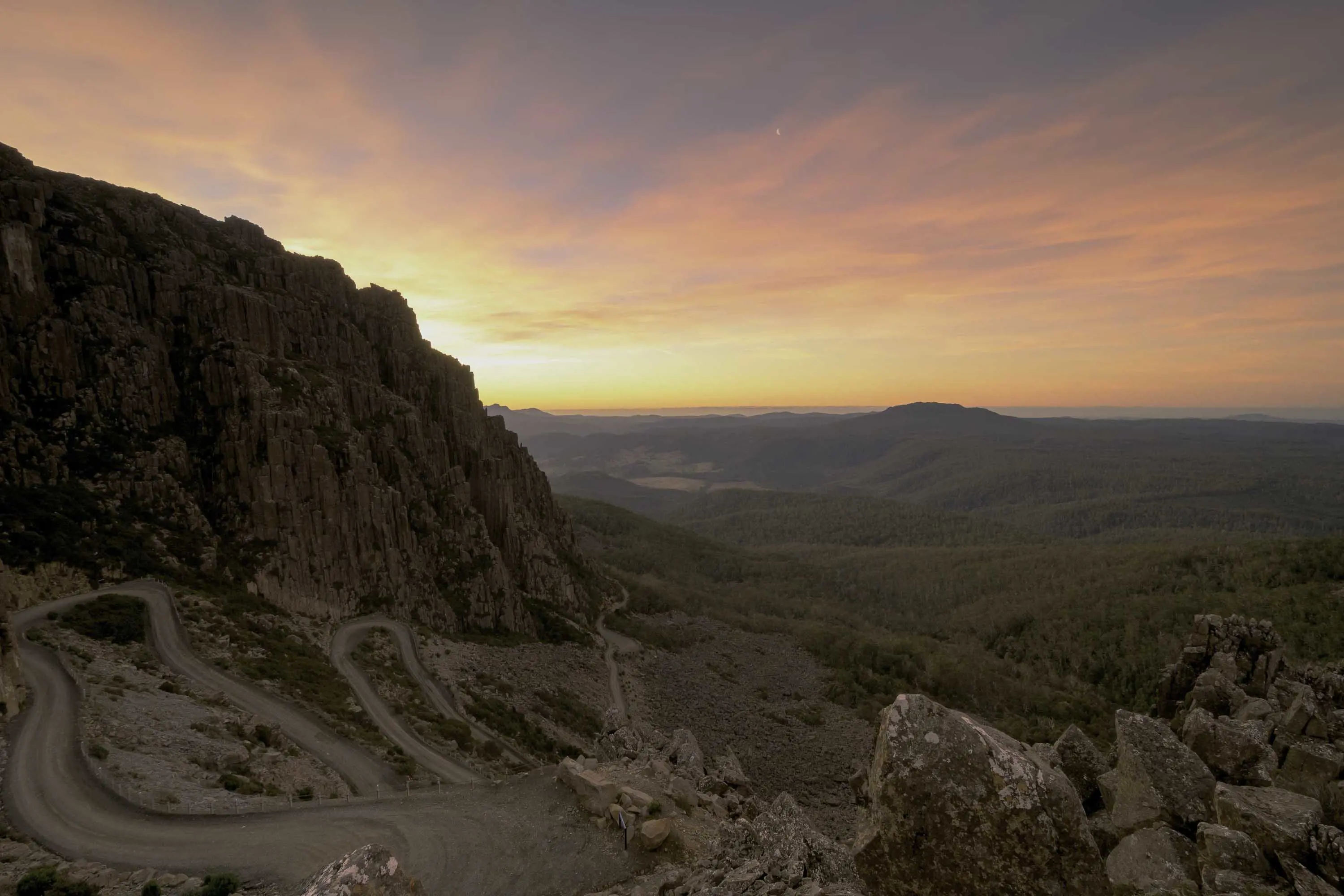 A sunset, seen from the top of a winding mountain pass, lights up clouds in orange, pink and yellow.