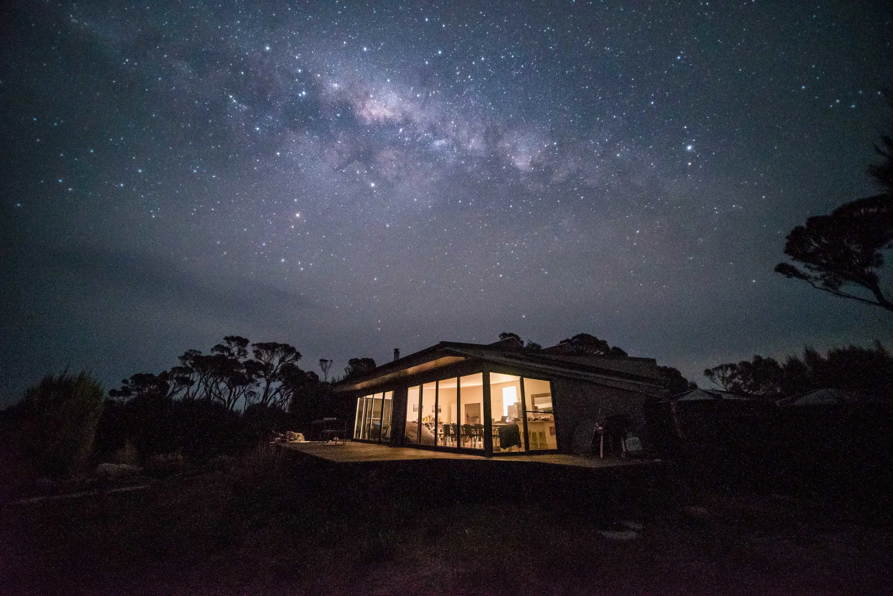 A modern house with tall windows at night with the mily way clearly visible in the night sky above.