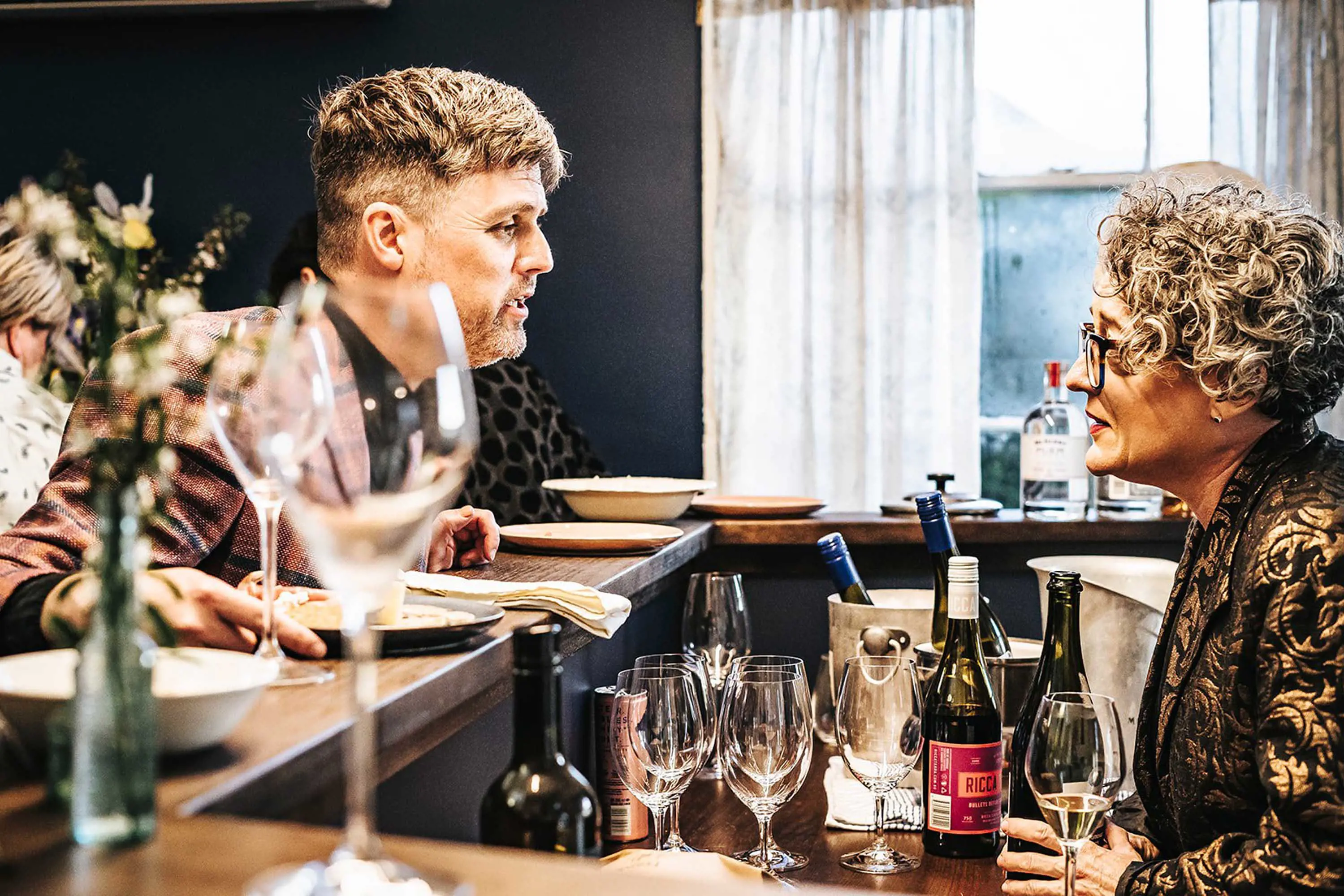 A man leans over a bar to talk with a woman who is serving wine from bottles on the counter.