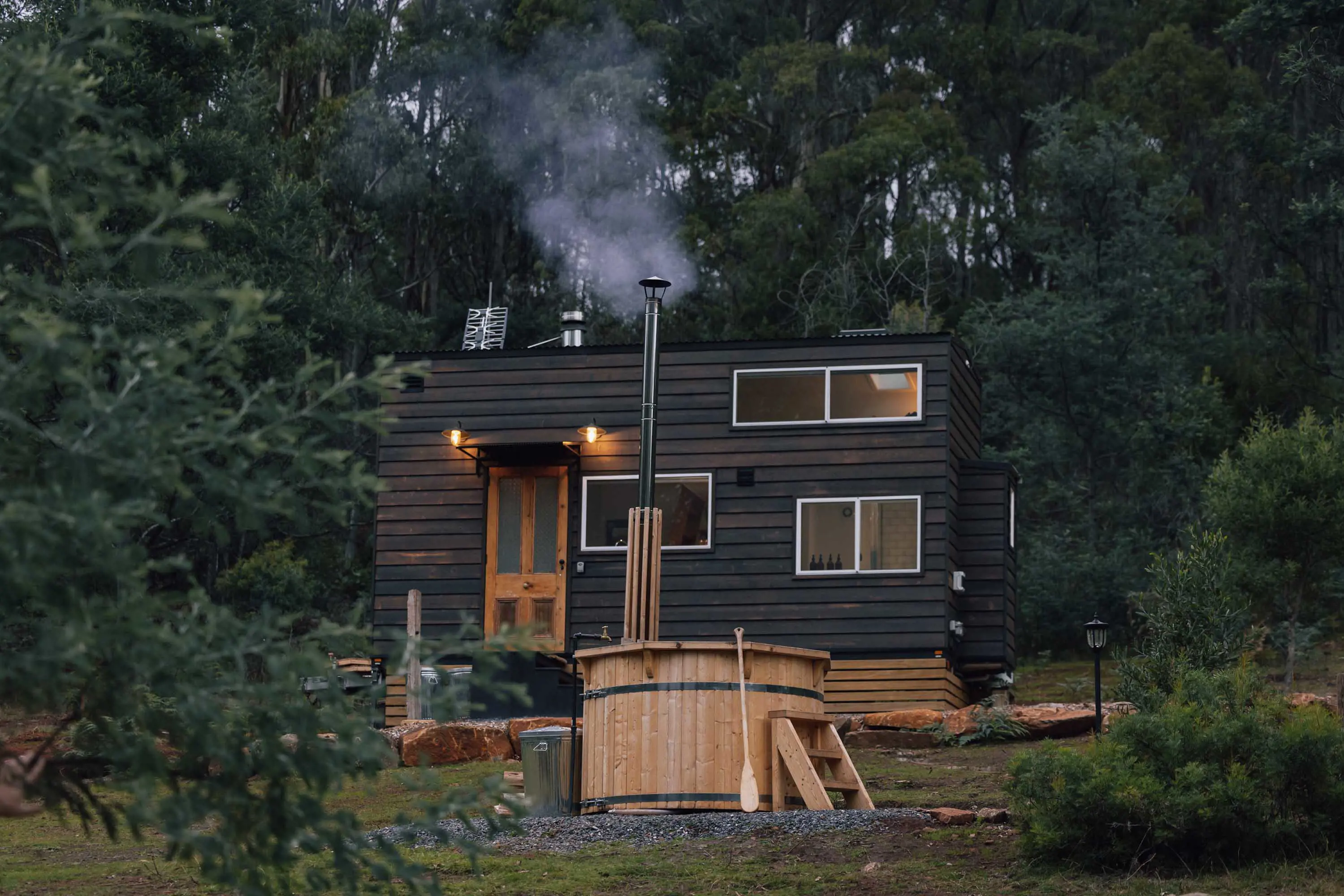 A wooden two-story cabin with dark brown, shou sugi ban charred cladding stands in a forest. A large outdoor spa made of pine stands in the foreground.