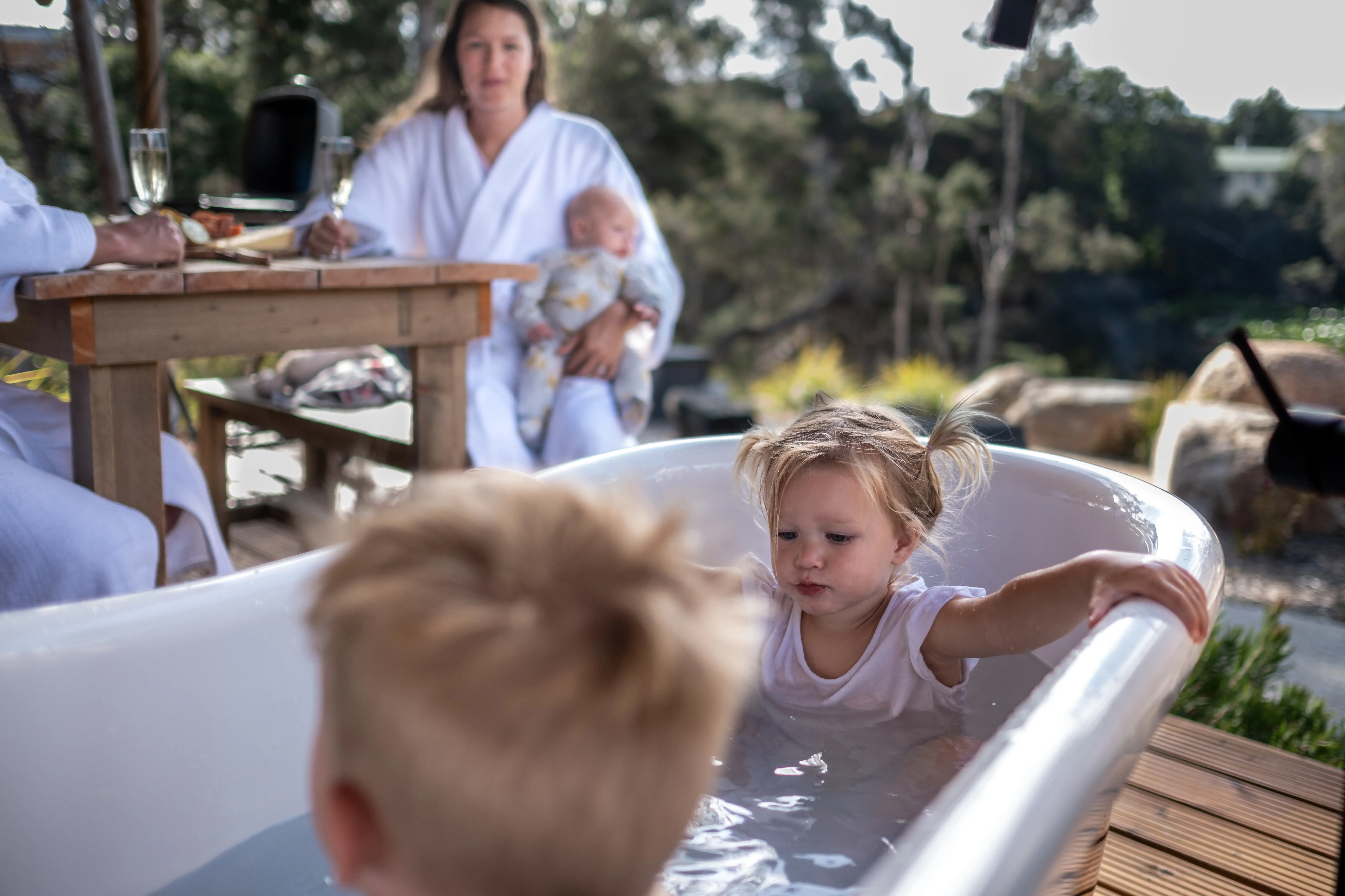 Two kids sit in a shallow bathtub on an outdoor deck, while two adults in white bathrobes sit watching them at a table.