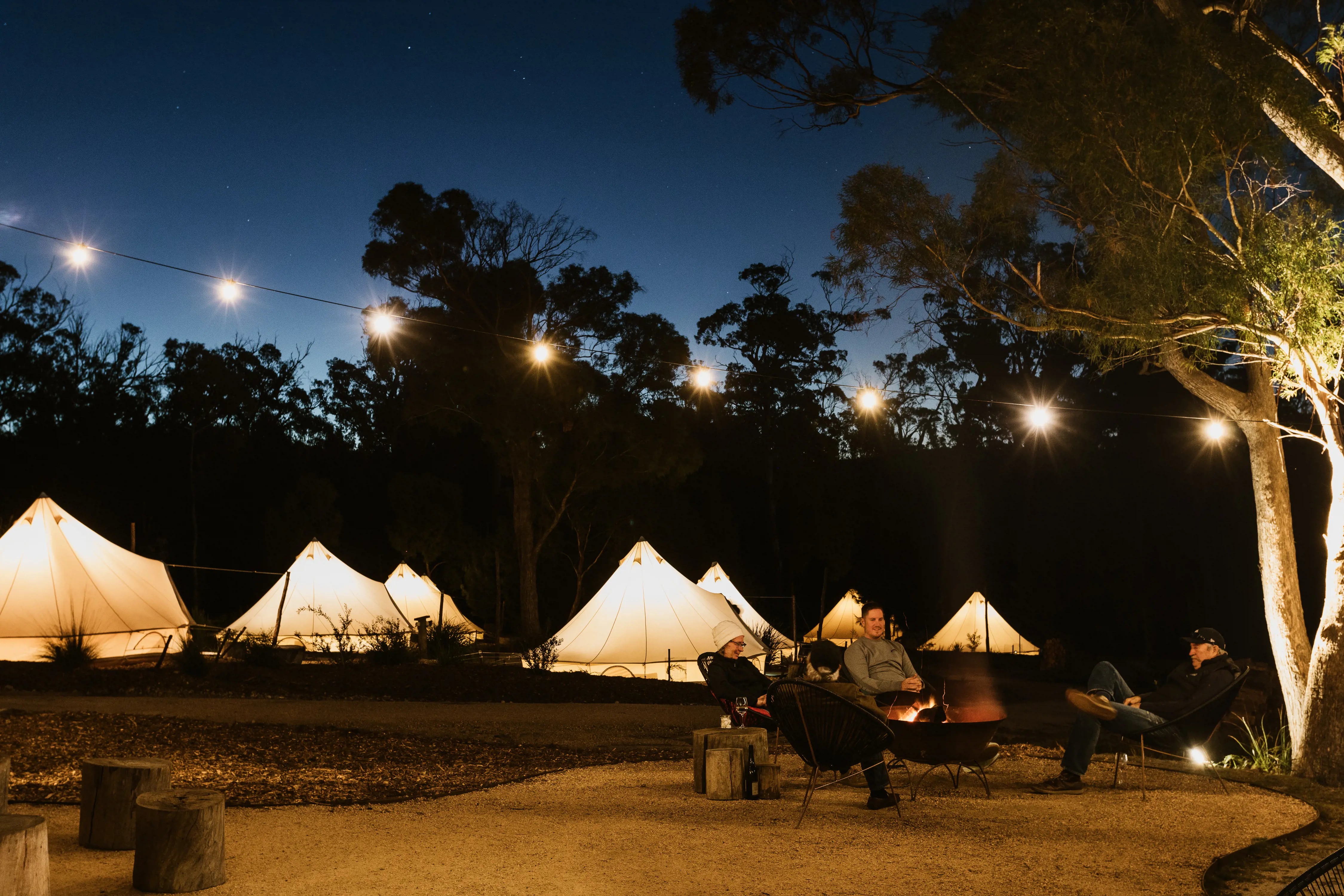 Several canvas tents lit up from the inside are dotted amidst the bush at dusk, with people sitting around a campfire under fairy lights strung between trees.