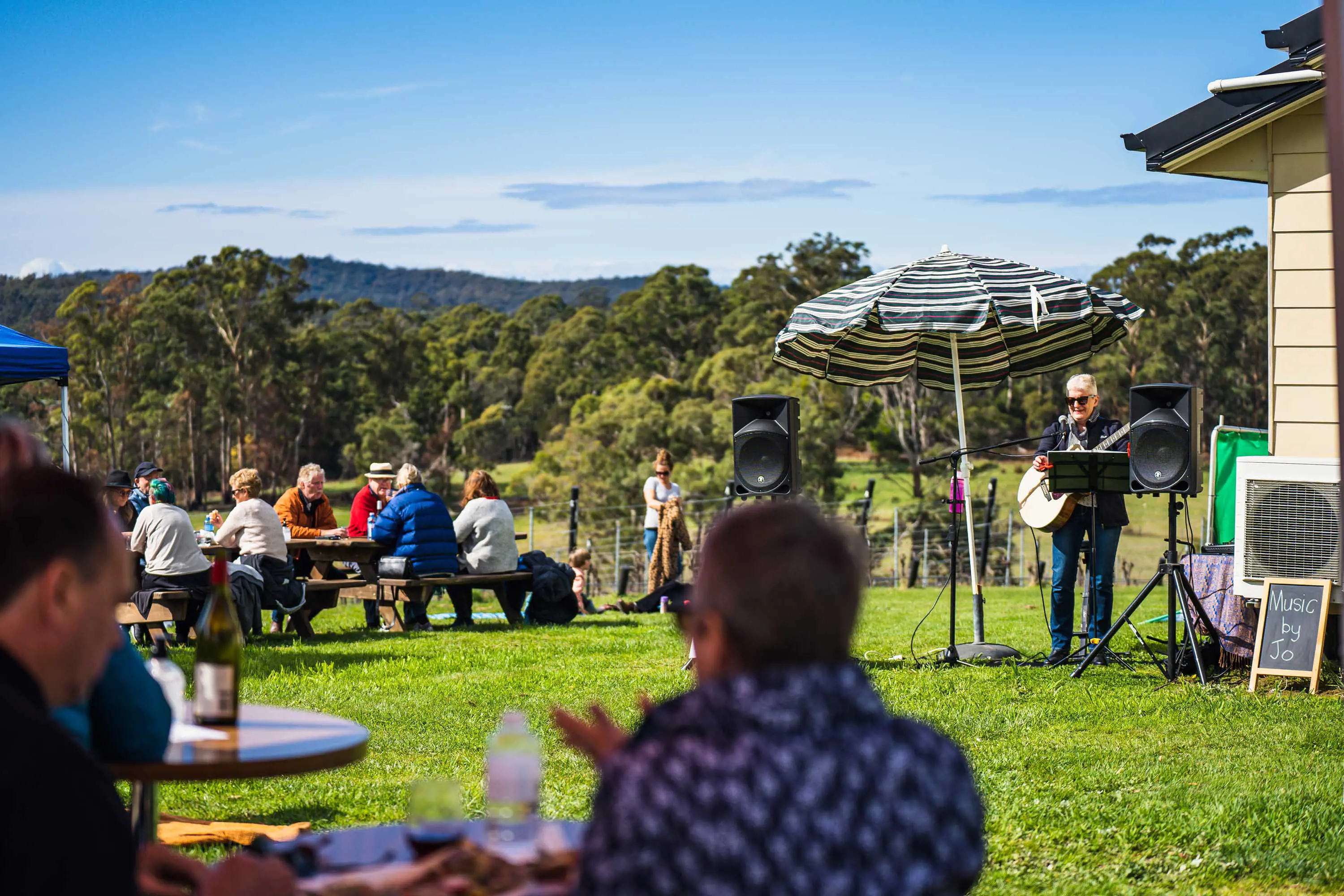 Groups of people sit at wooden tables under umbrellas in a grassy field next to a house. A musician plays a guitar and entertains.