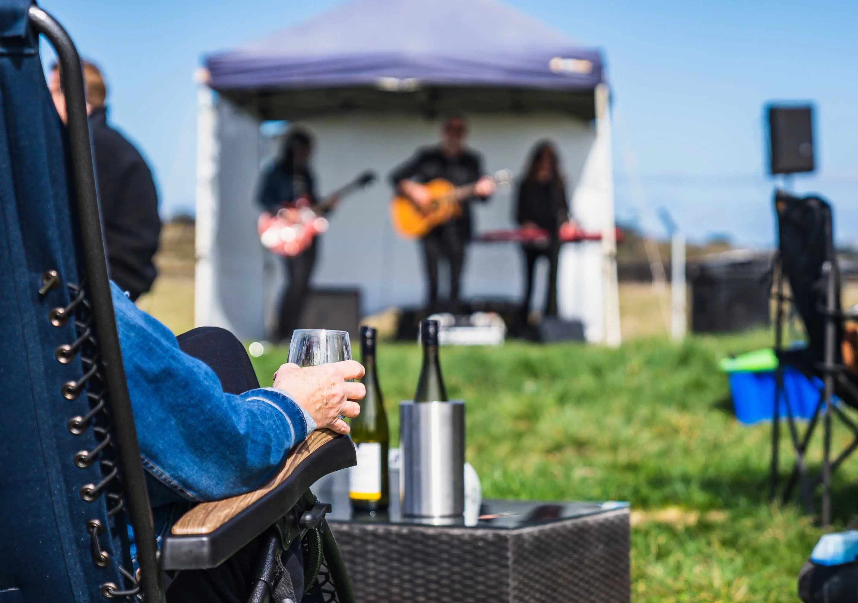 People drink wine and relax in recliners in front of a small, covered stage, where a three-piece band plays music.