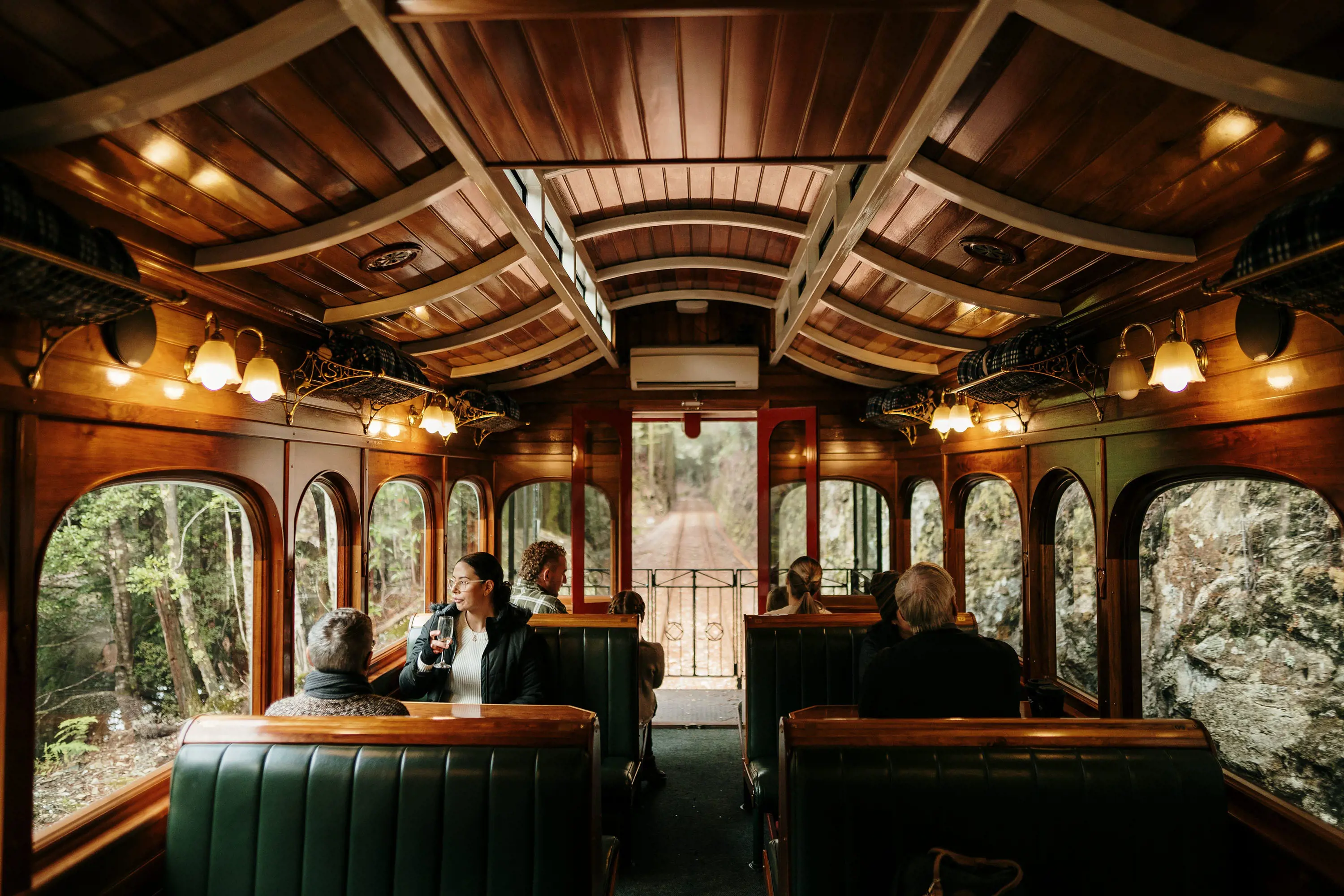 The gleaming polished wooden interior of a heritage train carriage, with booths either side of the aisle and people seated watching the forest go by.