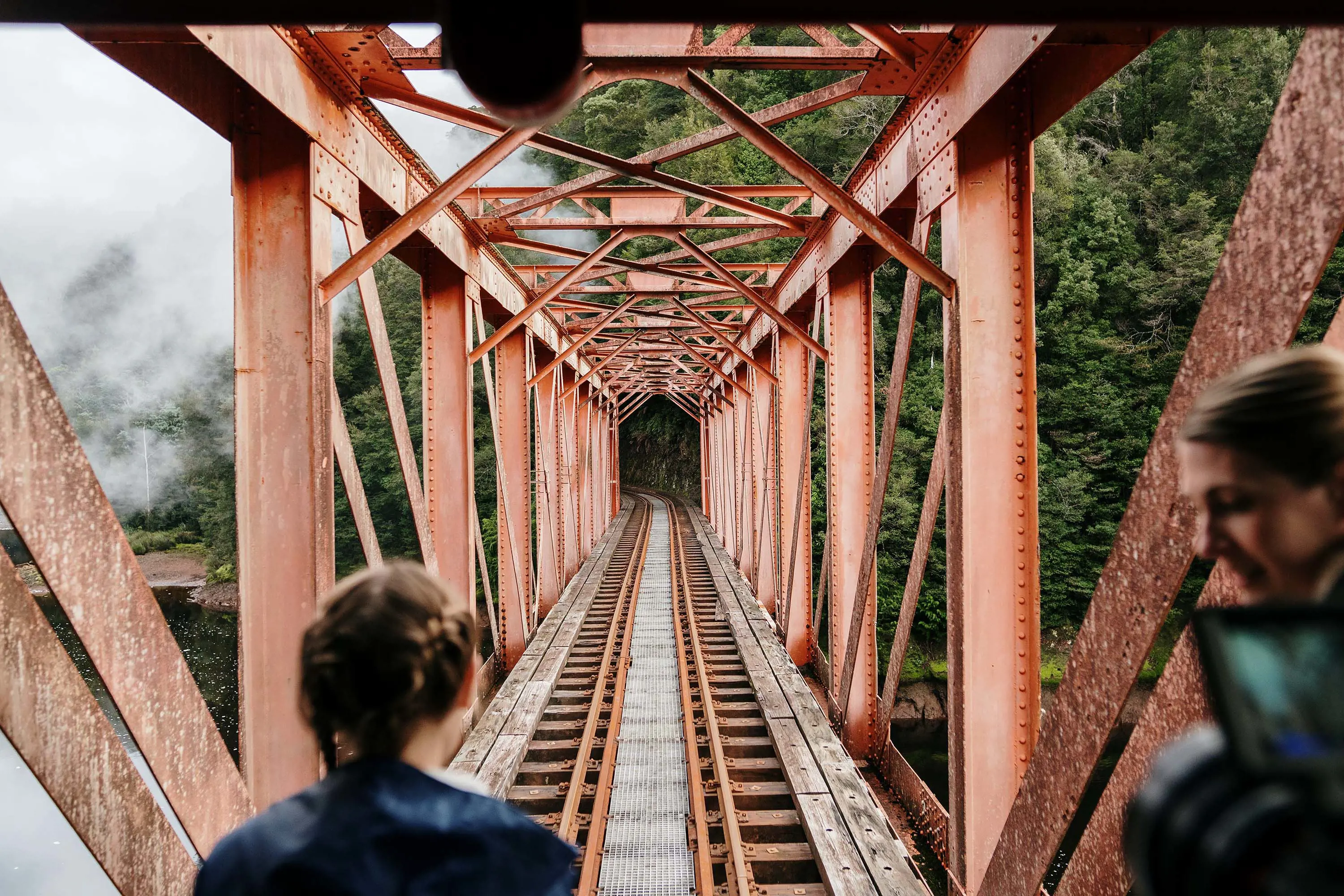 Two people stand in the middle of an old railway line, enclosed in a structure of faded red steel.