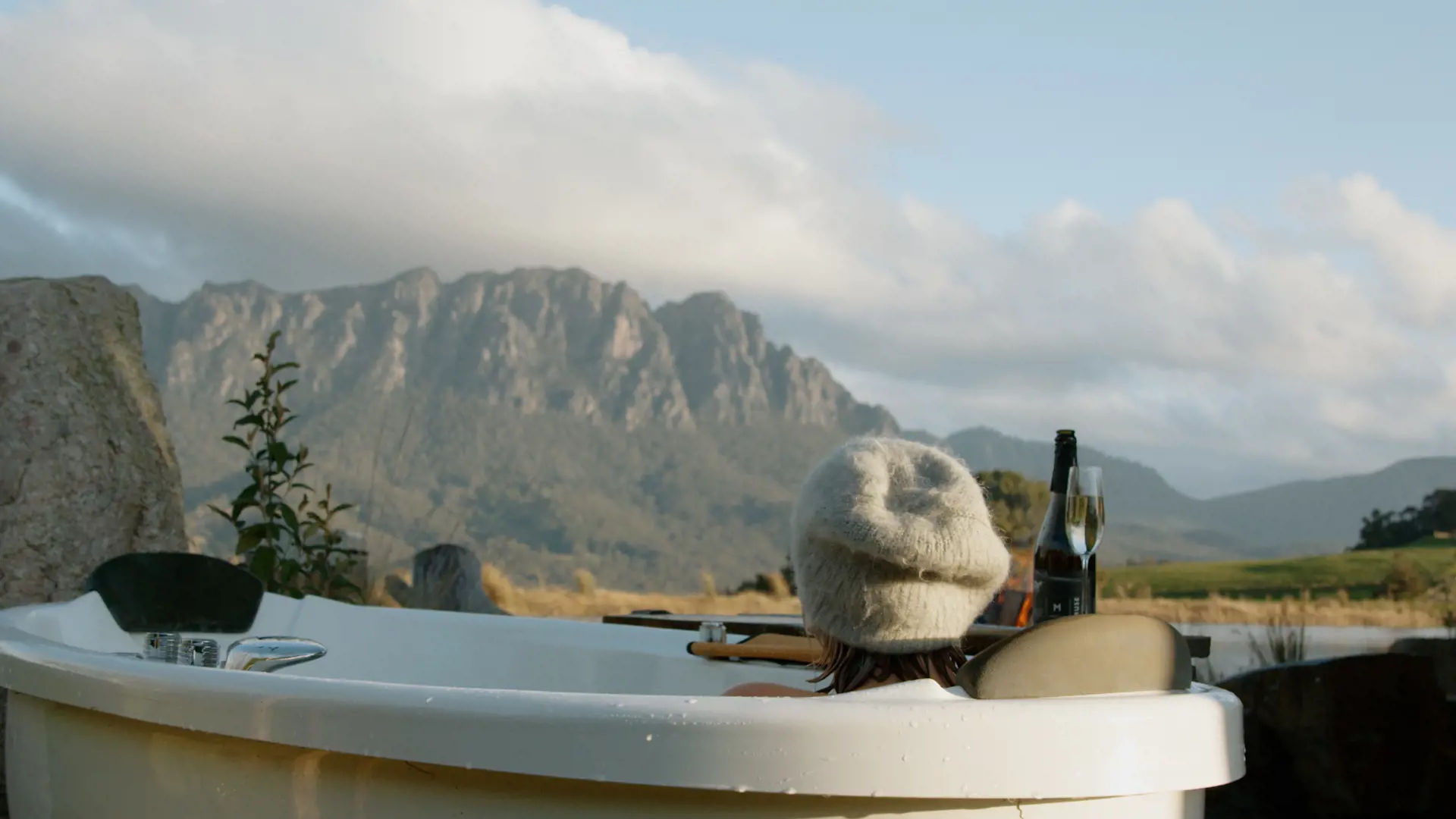 A woman wearing a white, woolen beanie reclines in a large white bath sitting on farmland facing tall, rocky mountain peaks in the distance.