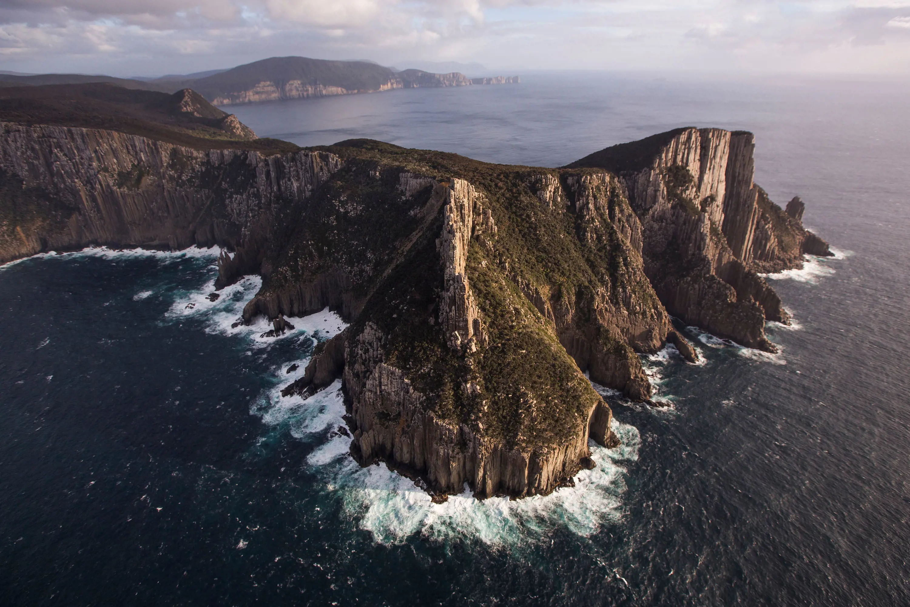 An aerial view of a rugged coastline of land jutting into the ocean, with sharp cliffs and crashing waves.