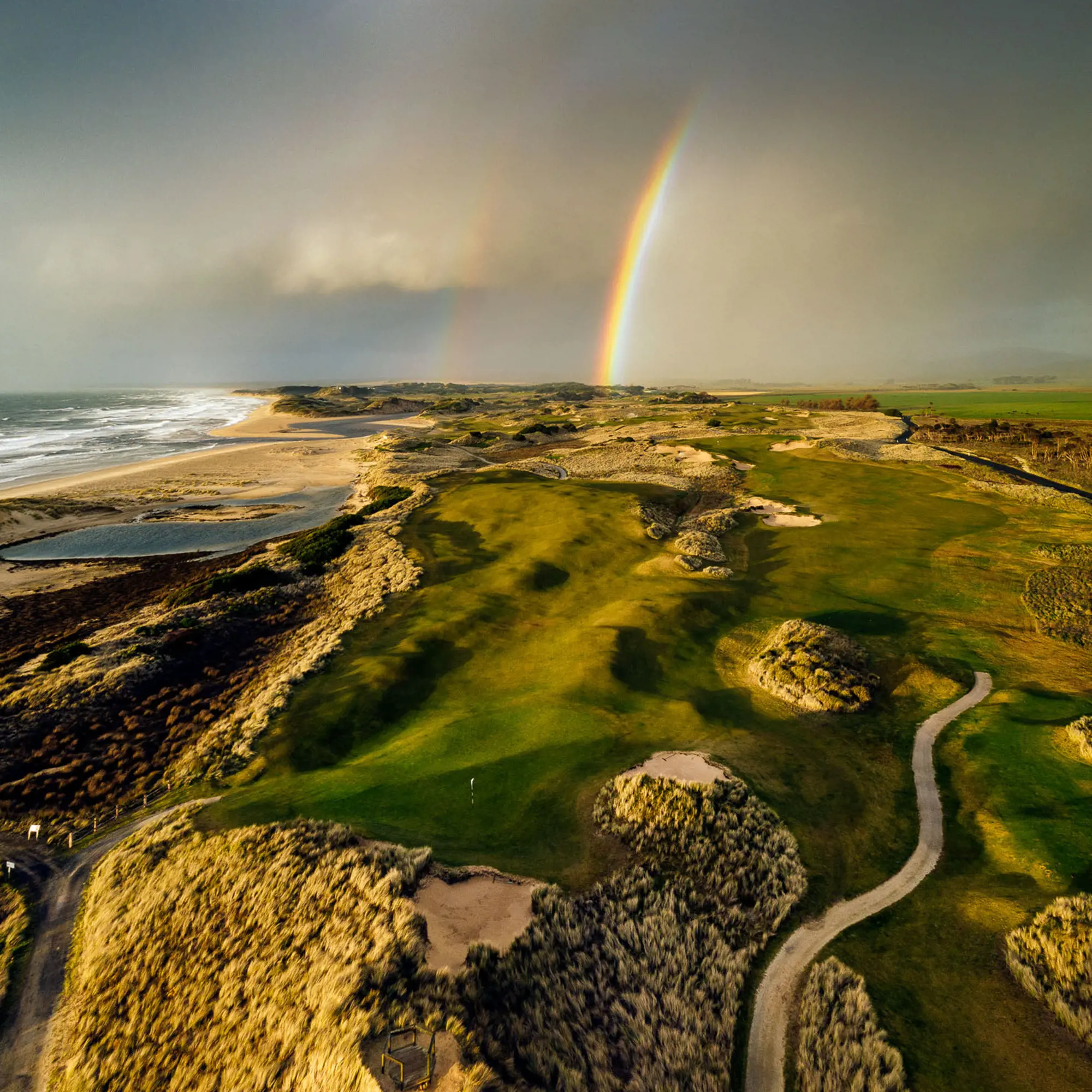 A coastal golf course winds around large sand dunes. A rainbow is visible in the sky.