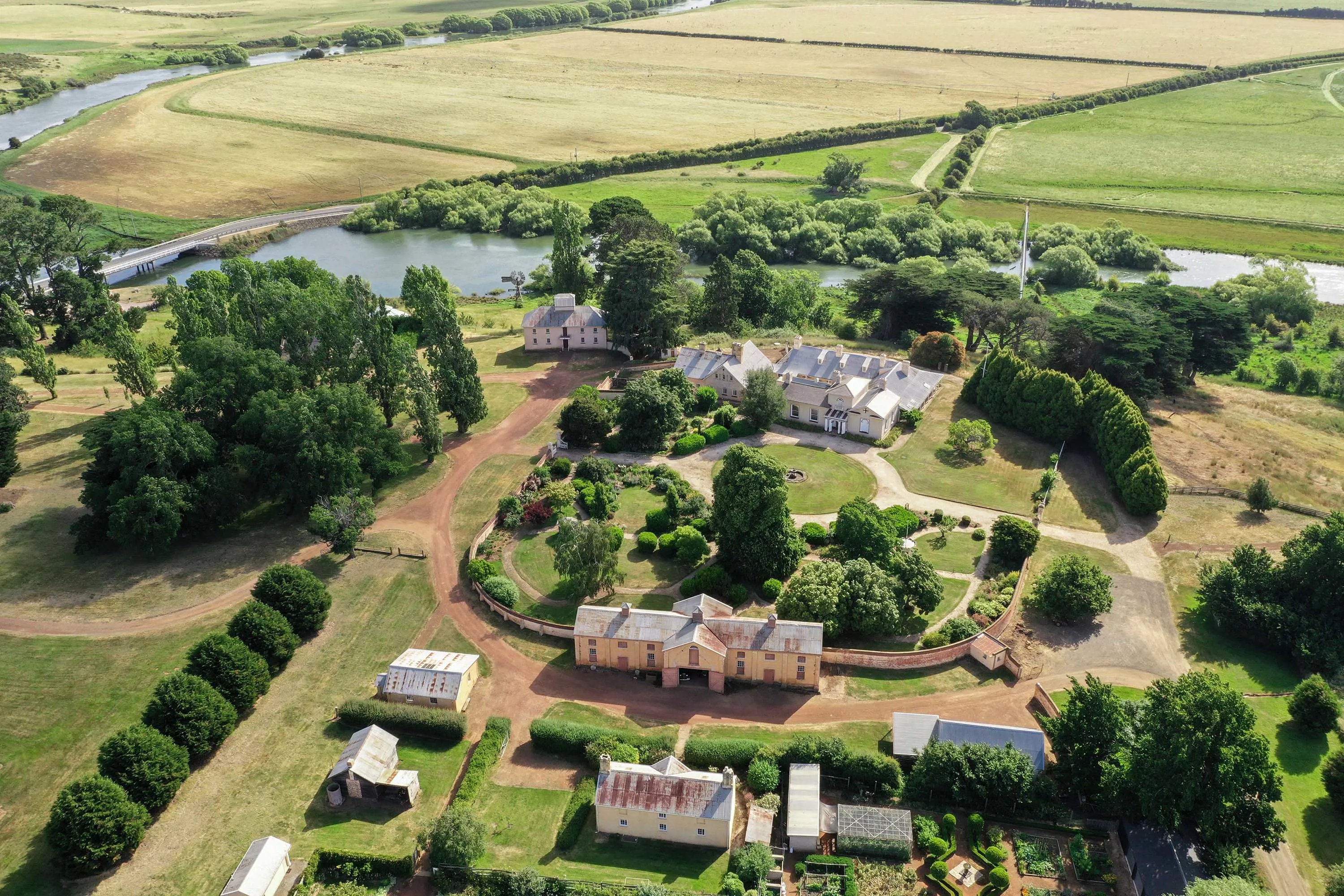 An aerial view of estate grounds, with manicured hedges, sweeping gravel driveways and beautiful old buildings, situated next to a river.