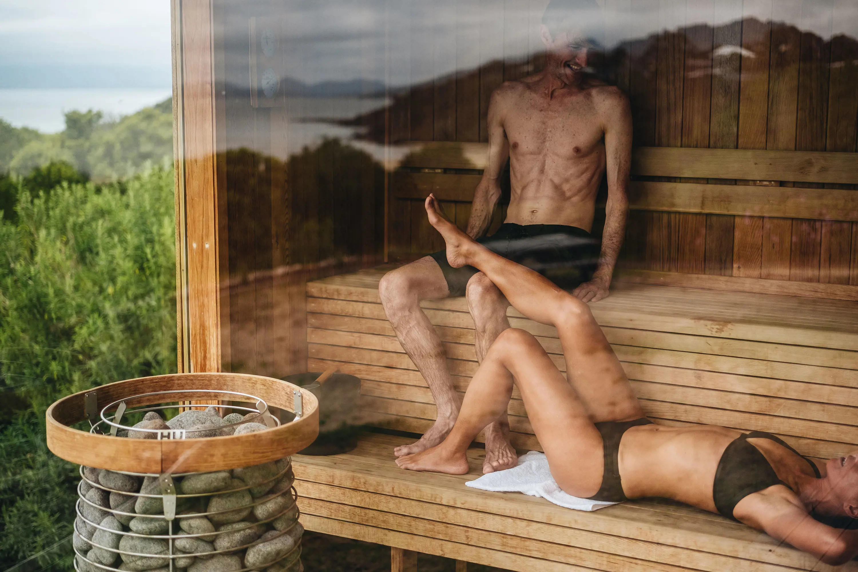 Two people sit in a sauna with views of bushlands near the beach through a large glass window.