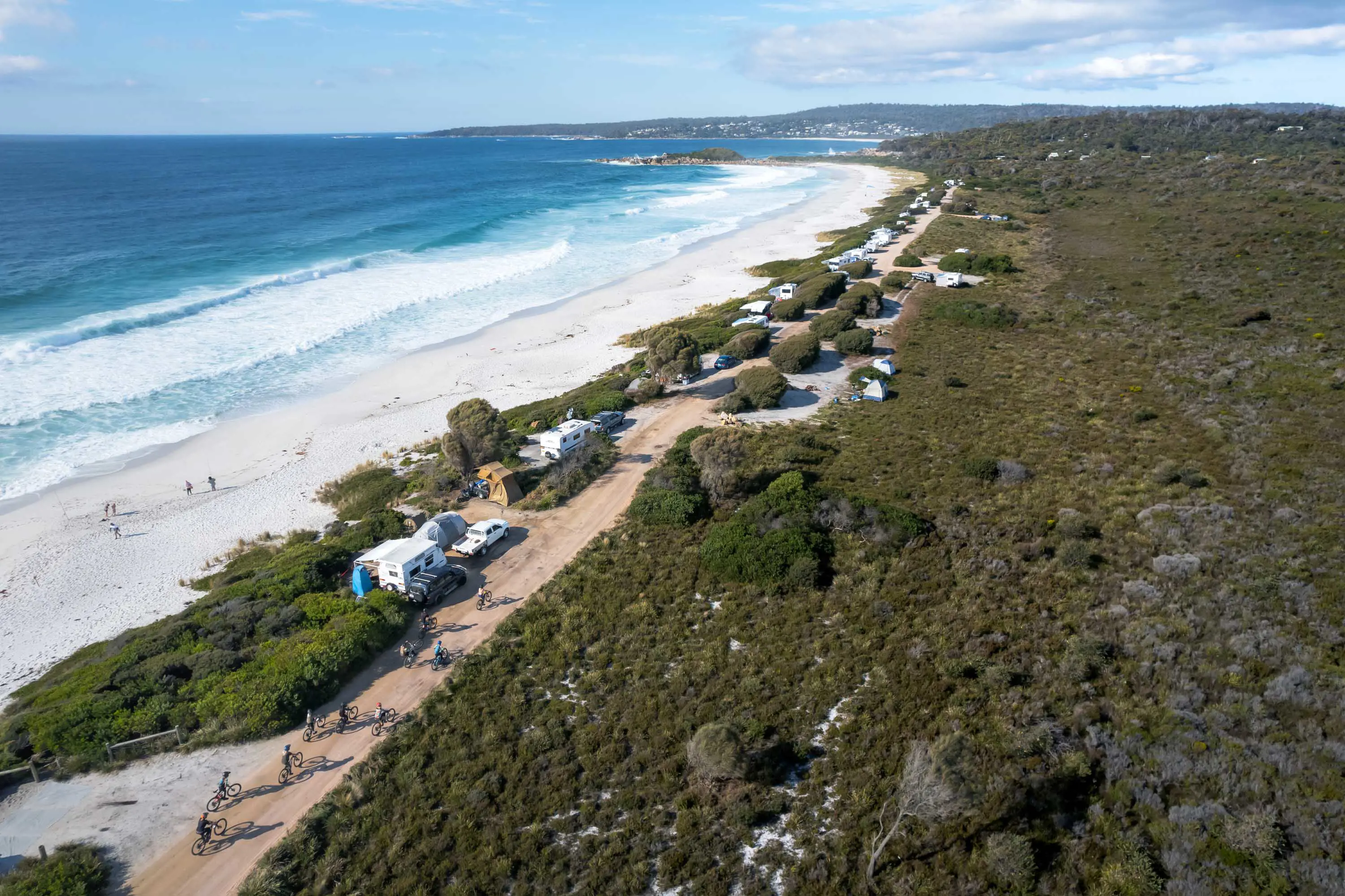 Caravans and tents stand near bushland looking over white sands and blue waters of the ocean.