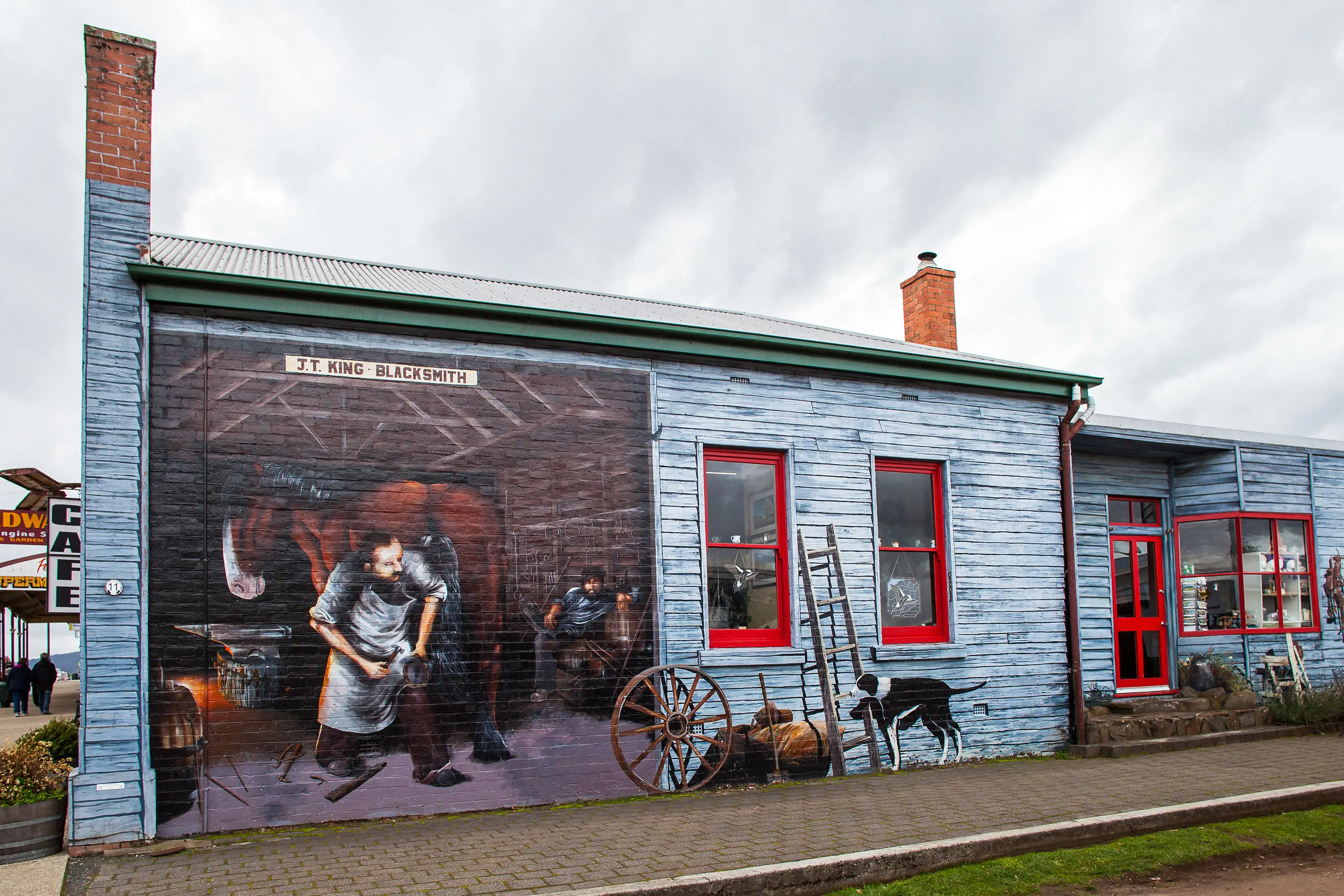 The side of a brick building is painted intricately to appear like a blue weatherboard building, with scenes of a blacksmith's inside.