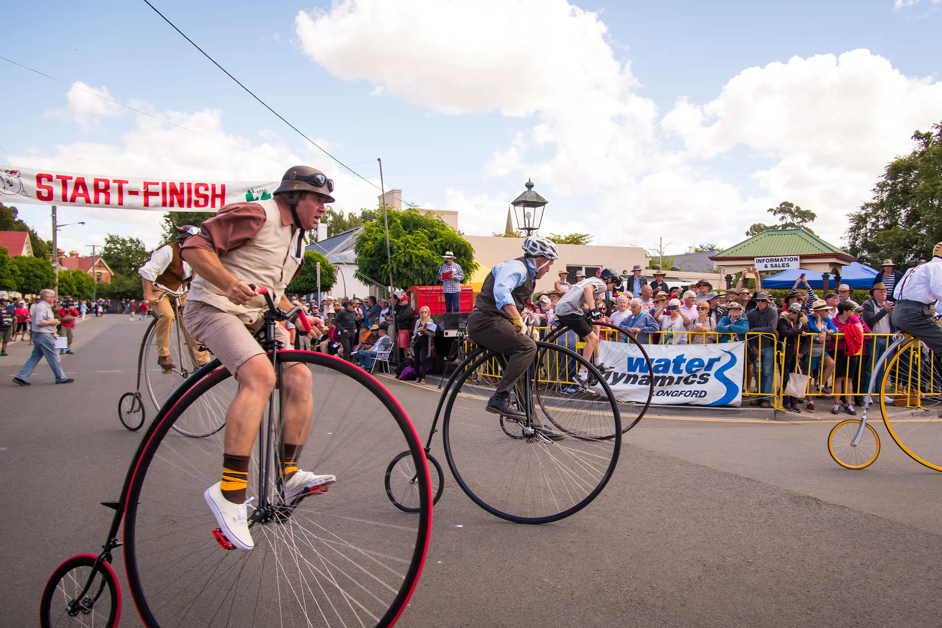 Men dressed smartly riding penny farthing bicycles on a wide road are cheered on by crowds of people behind a yellow barrier.