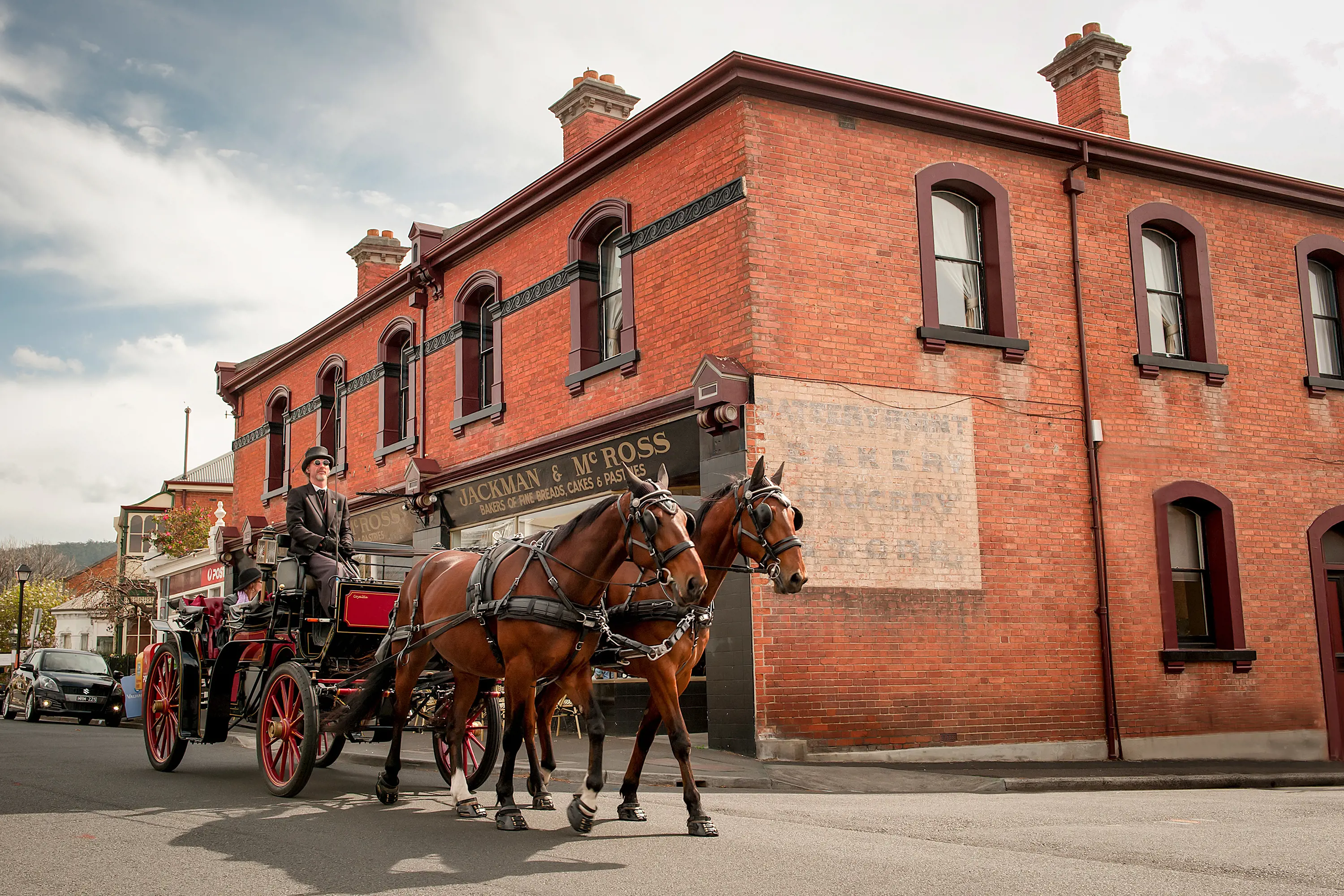 Two horses pull a red and black traditional carriage down a street. A smartly dressed man in a top hat sits in front driving the carriage.