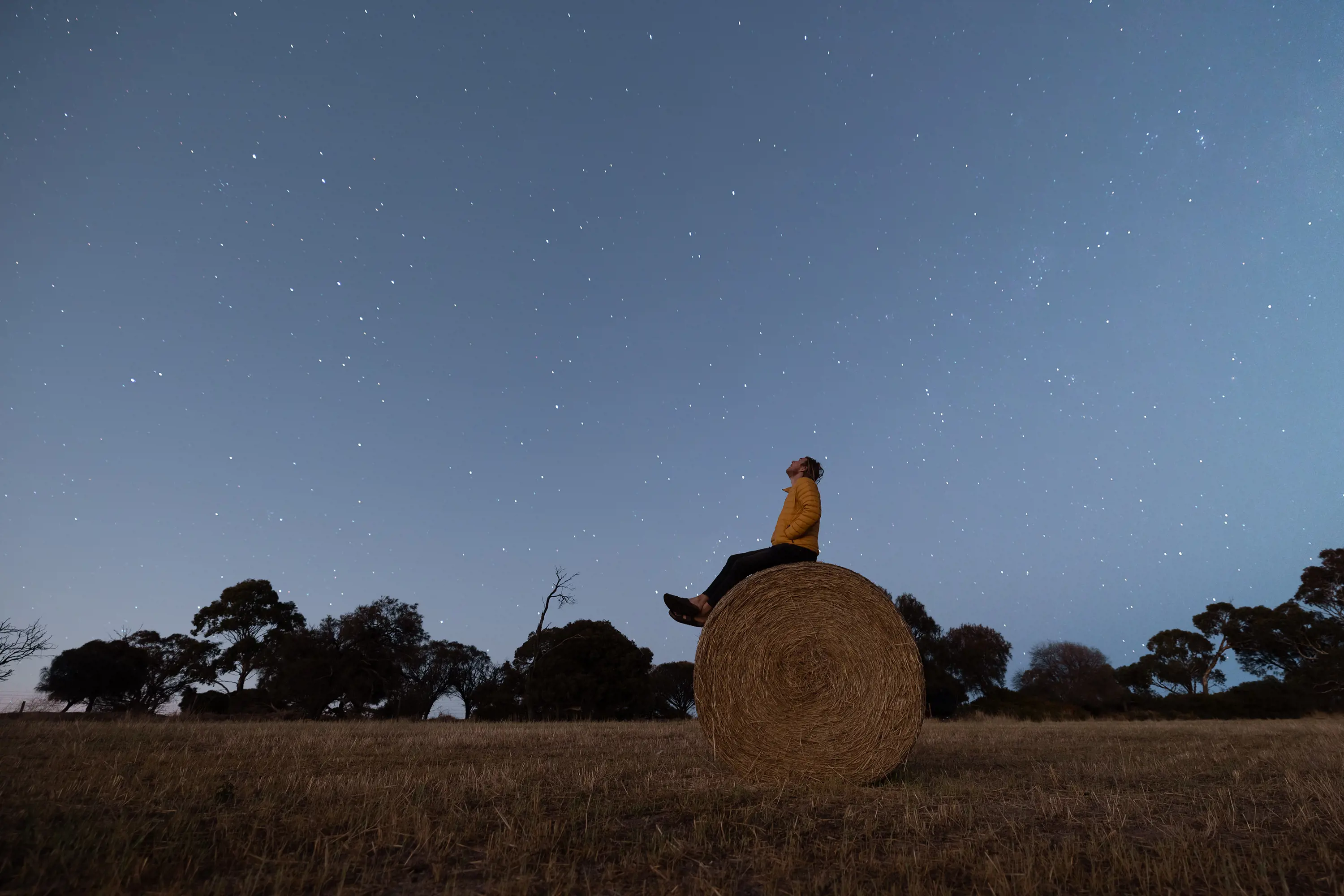 A person sits on a large round hay bale in the middle of a field, looking up into the night sky full of bright stars.