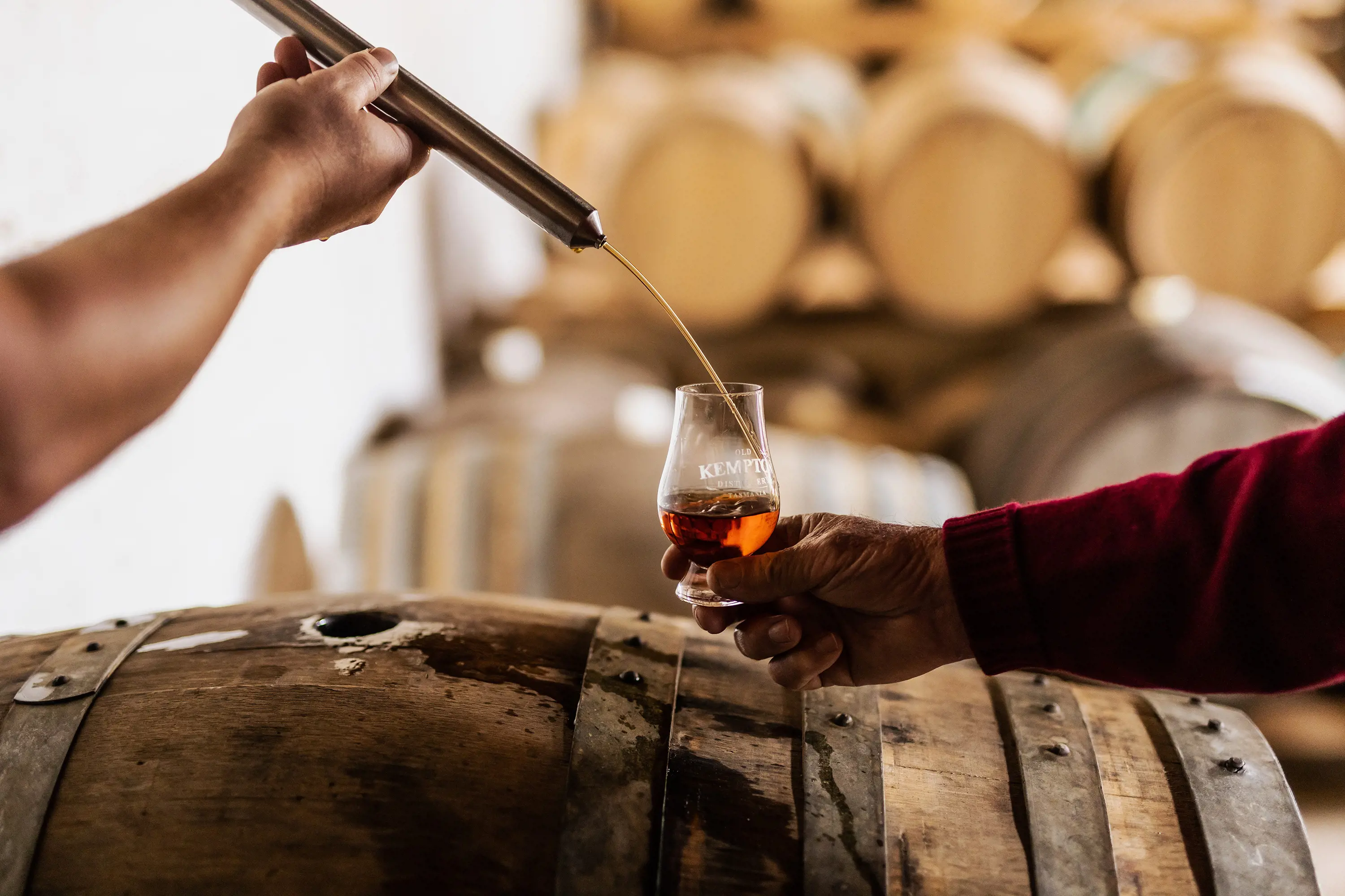 A person pours whisky from a metal tube into a shapely glass, over a barrel.