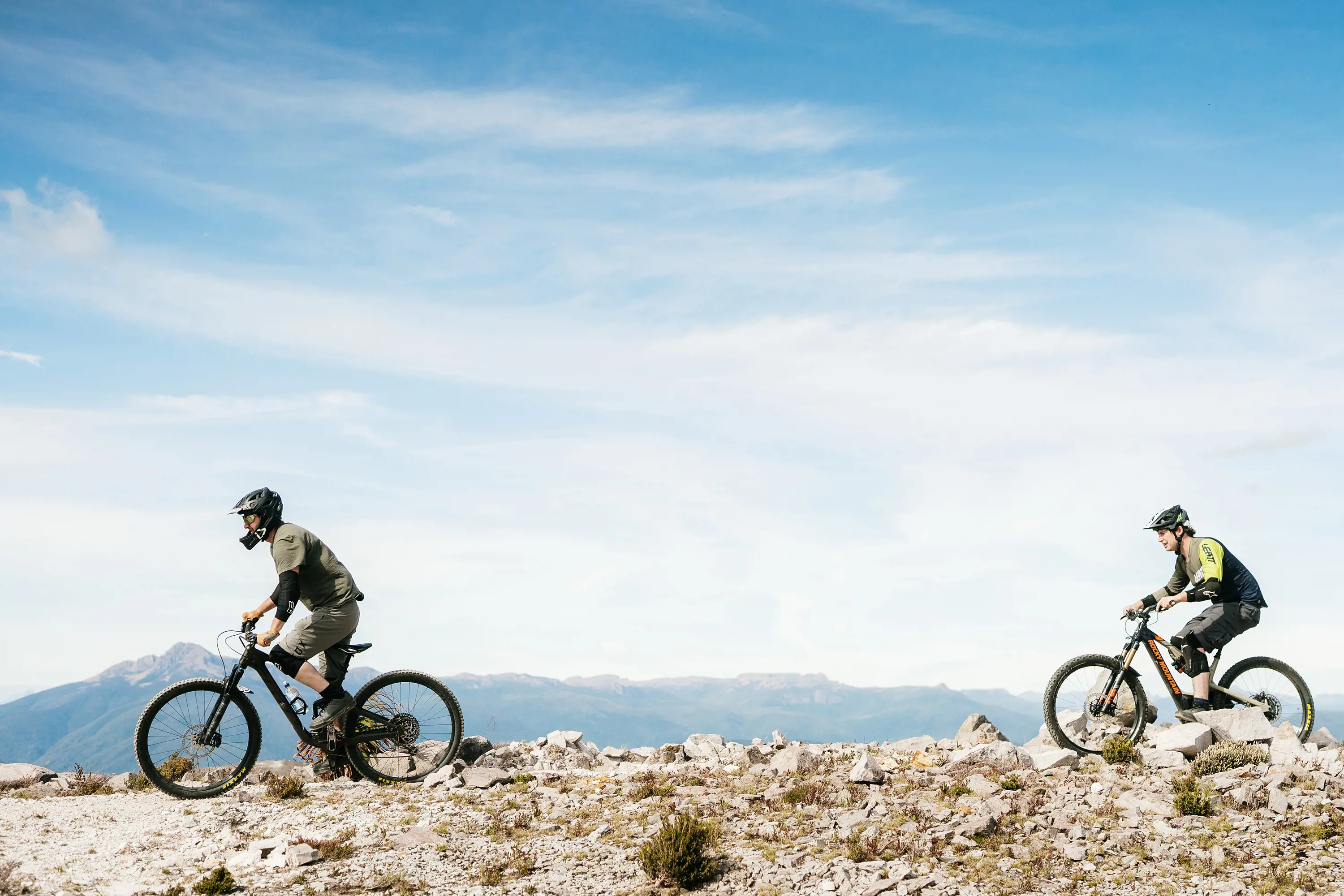 Two people on mountain bikes ride across a flat, open terrain. Behind them the path seems to drop off into nowhere, with mountains rising behind.