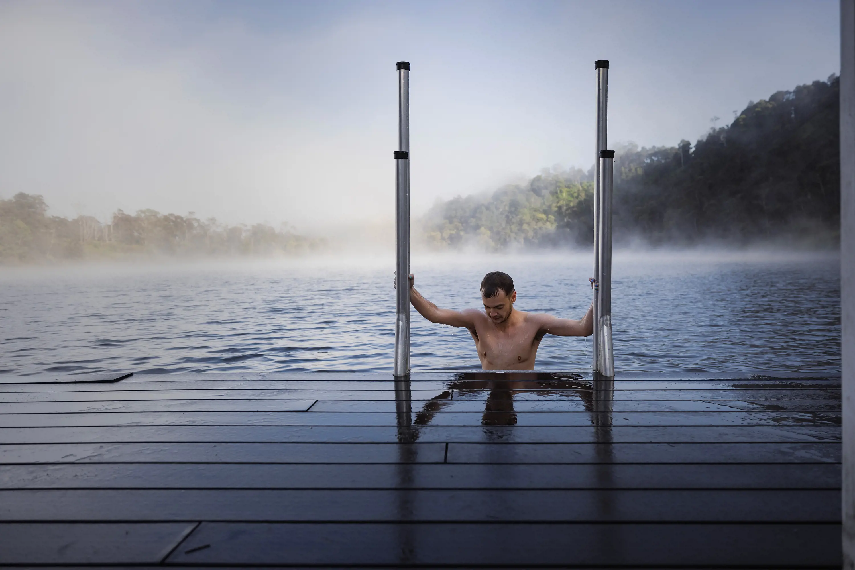 A man climbs from foggy lake waters onto a wet wooden deck.