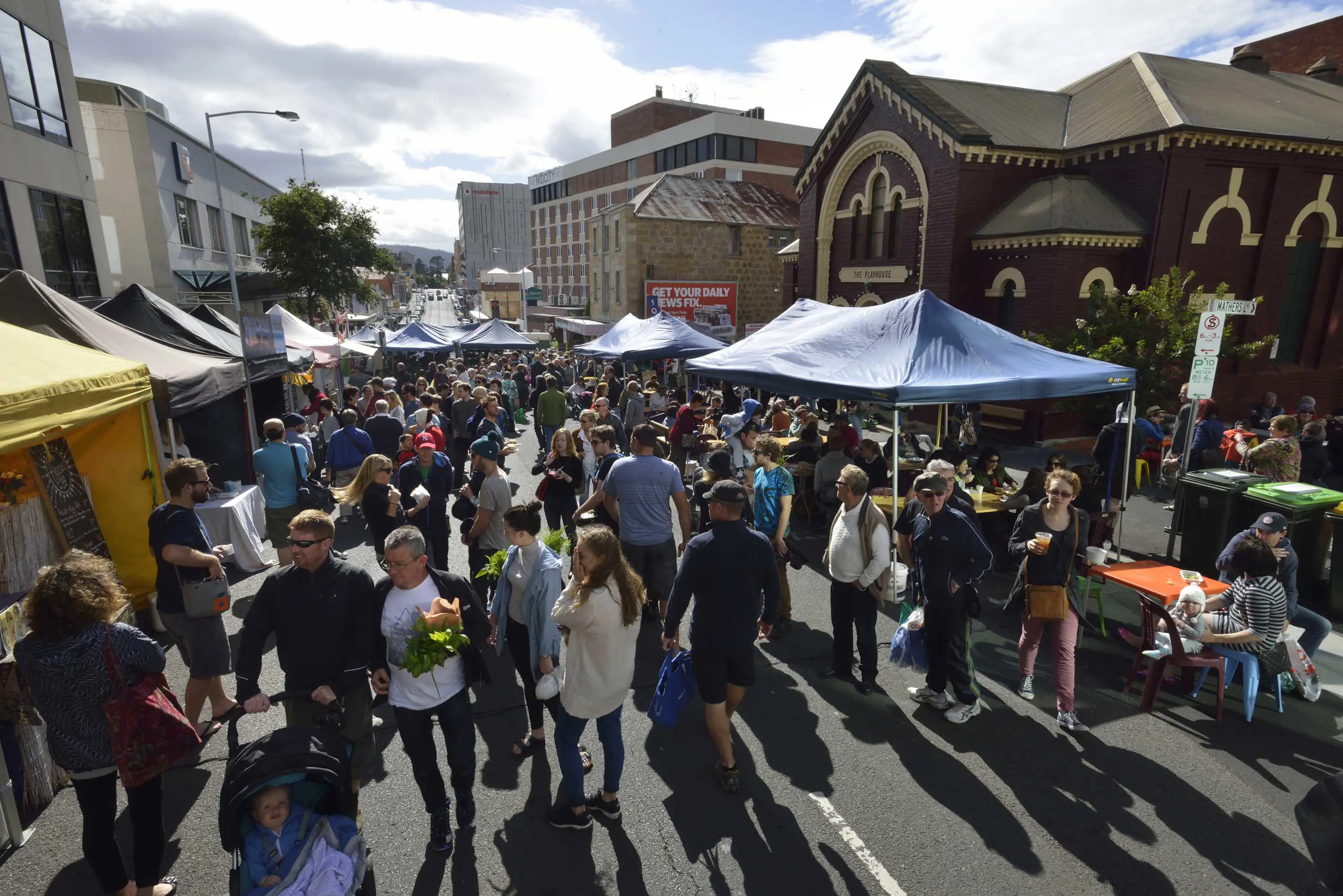 A large gathering of people walk between stalls placed in front of buildings on a closed street on a sunny day.  