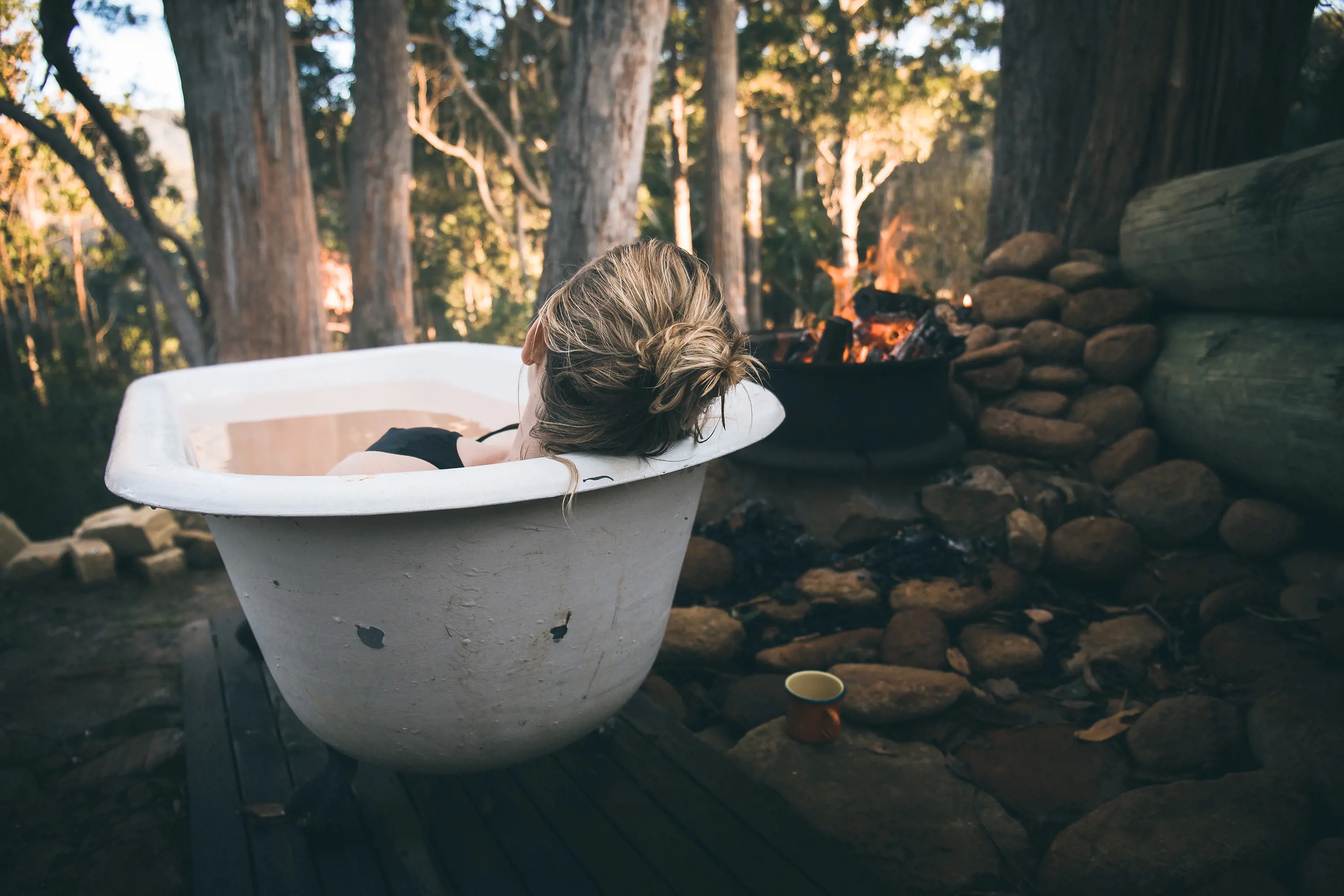 A woman relaxing in an outdoor bathtub looking out to the bush. Beside her, a firepit is lit and roaring.