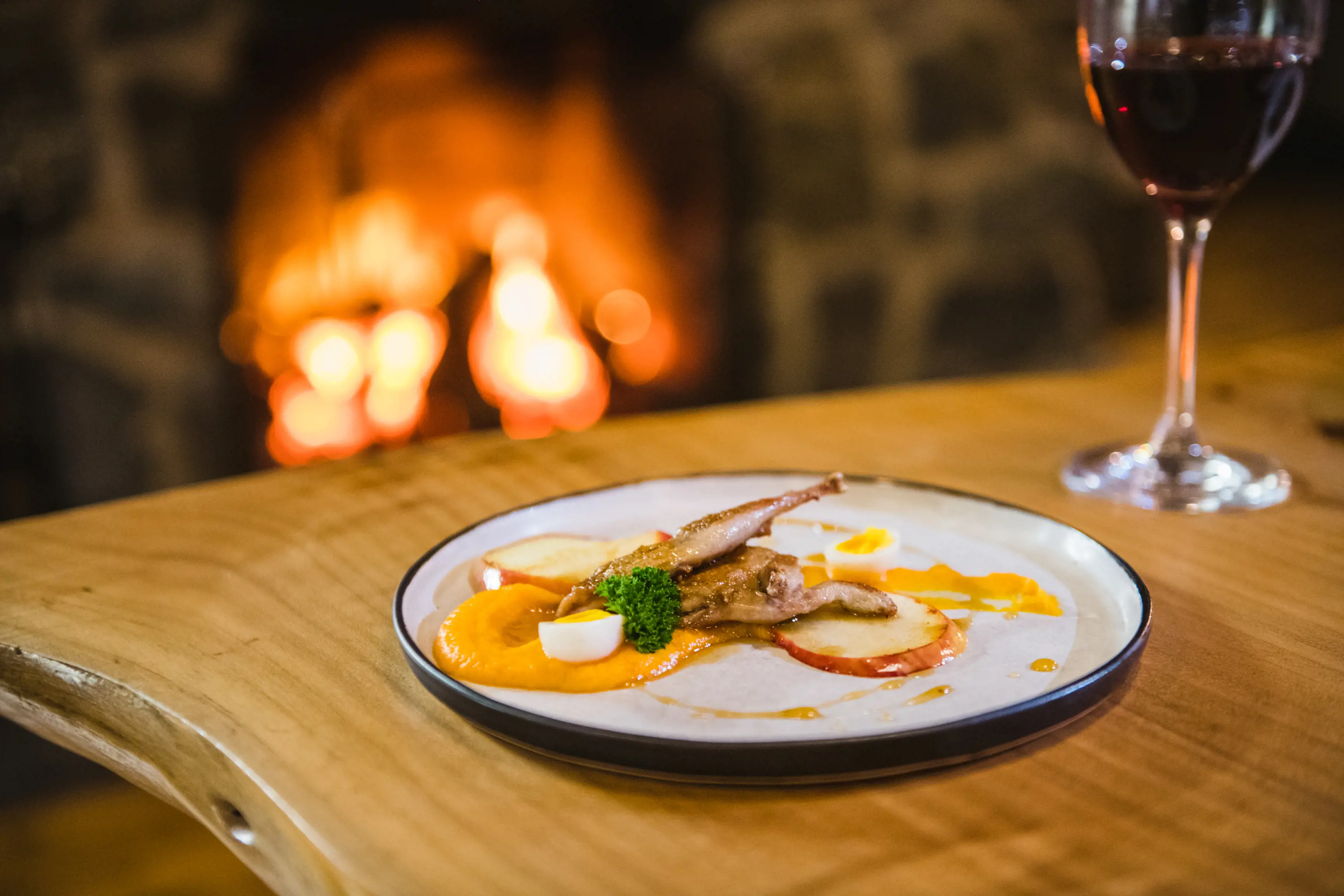 A plate of food and glass of red wine sit on a polished wooden table. A fireplace is roaring in the background.