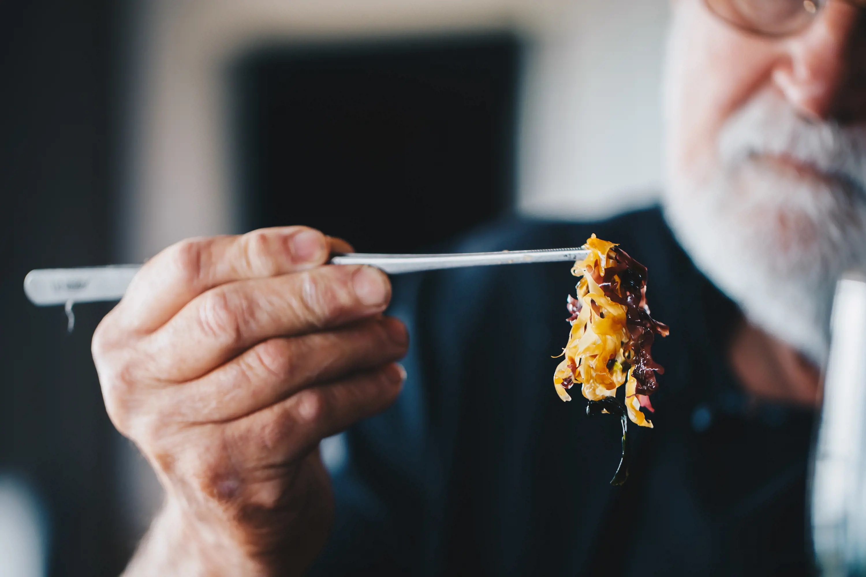 A man with a short grey beard uses long cooking tweezers to hold a small piece of yellow and brown kelp.