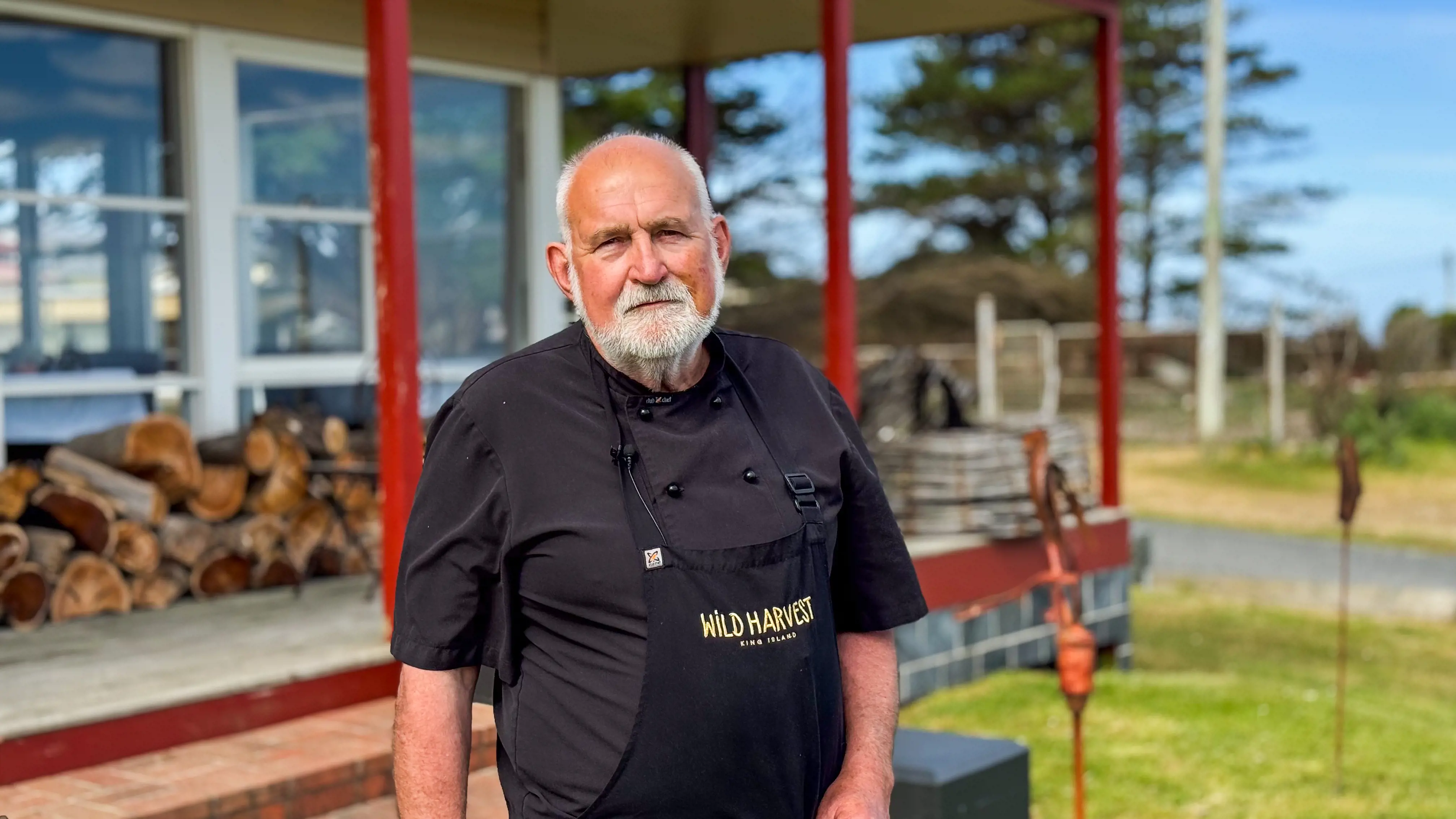 A grey bearded man wearing a black t-shirt and apron stands on the grass in front of a building. The walls are all glass and a pile of firewood sits on the deck outside.