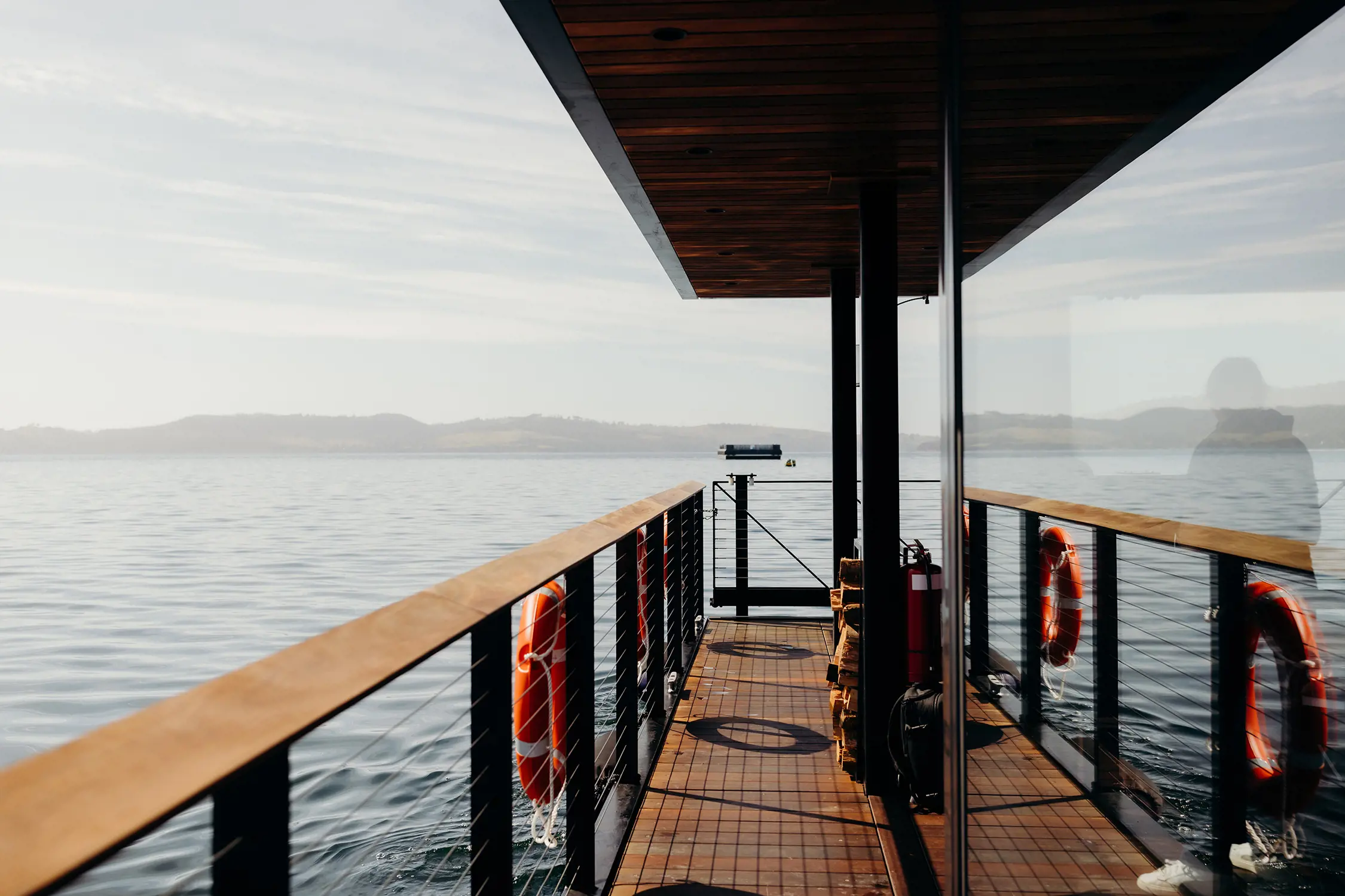 Onboard a floating sauna boat. The water beneath is dark and rippled. A wooden walkway wraps around the glass-encased sauna space, with orange lifebuoys hanging off the railing. The scene is reflected in the smooth glass wall.