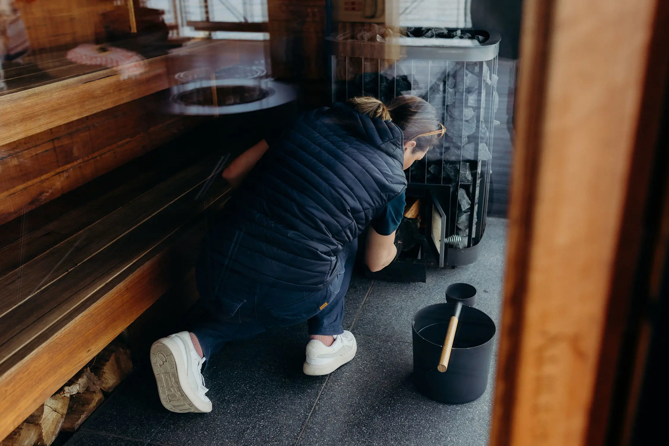 Inside the wooden panelled sauna, a woman crouches down, feeding wood into the tall sauna stove.