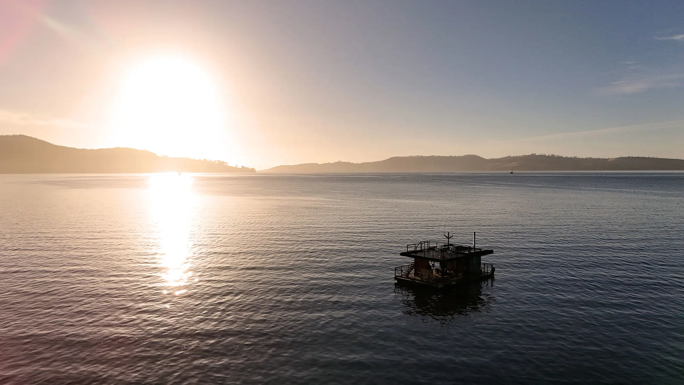 A rectangular pontoon-style boat, with a wraparound walkway and top deck, floats peacefully in the water of a wide river. The sunset casts a glow across the sky and reflects off the dark water.