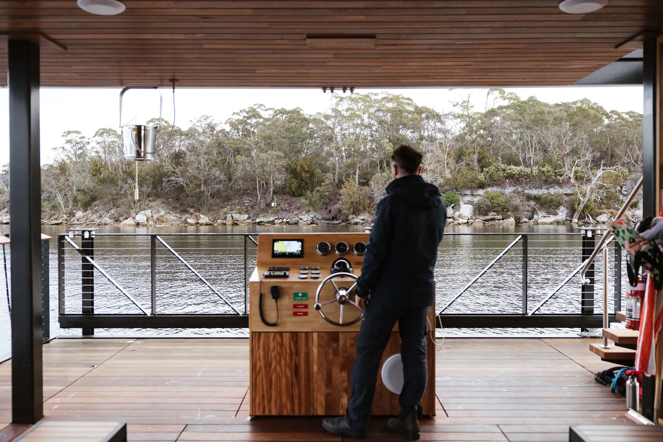 A wide open wooden deck with a small navigation panel island in the centre. A man stands at the metal steering wheel, looking out to the water and trees beyond.