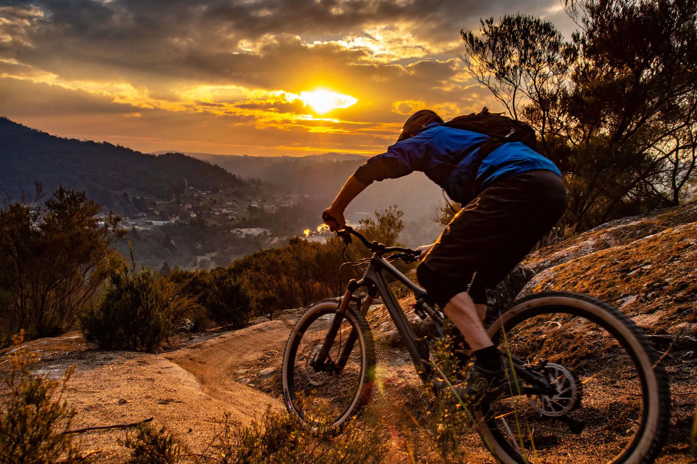 A person on a mountain bike is about to ride down a steep downhill track of sandy yellow dirt. In the distance, past a town and mountains, the sun peeks through some heavy grey clouds.