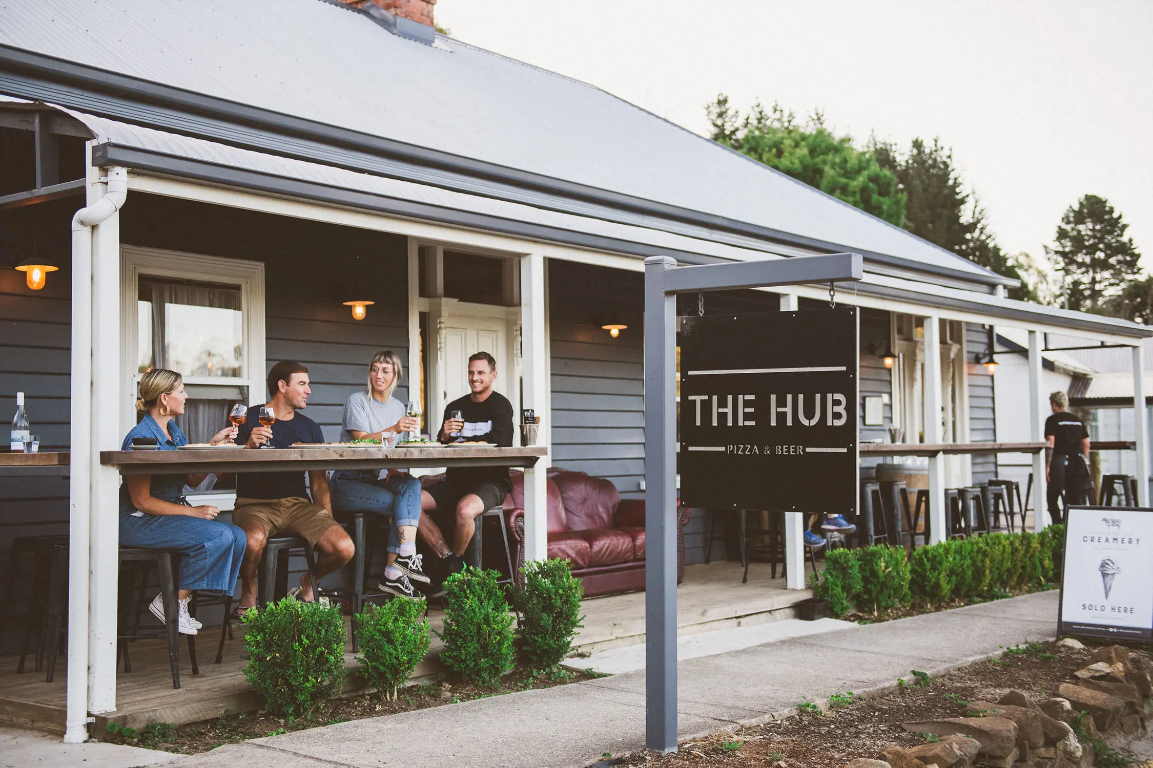 Outside a blue weatherboard building, four people sit at a bar table holding glasses of wine. A sign hanging from a signpost out the front says "THE HUB".