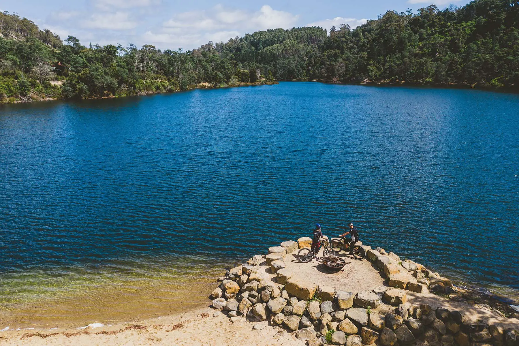 A richly blue lake surrounded by bush. On the shore, atop a rocky lookout structure, two people sit on bikes.