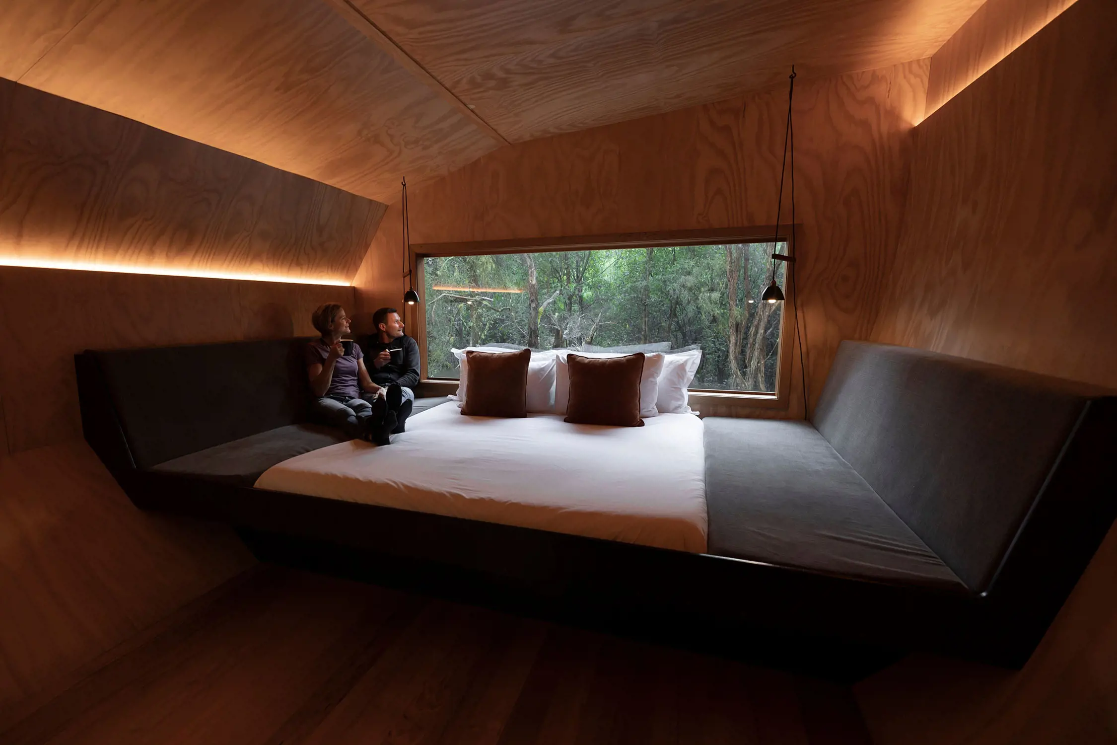 Inside a sleek, minimalist cabin, a large bed with crisp white linens sits against a natural wooden wall. Opposite the bed, a floor-to-ceiling window offers an immersive view of the surrounding dense forest, allowing natural light to fill the room. Two people sit on the bed, enjoying a moment of quiet relaxation