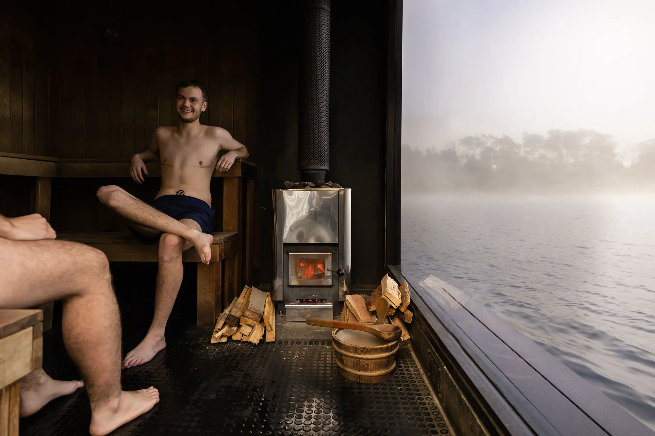 Inside a sauna, two people sit on wooden benches. The sauna stove is lit with stacks of wood next to it. Through the glass wall, a misty lake is visible.