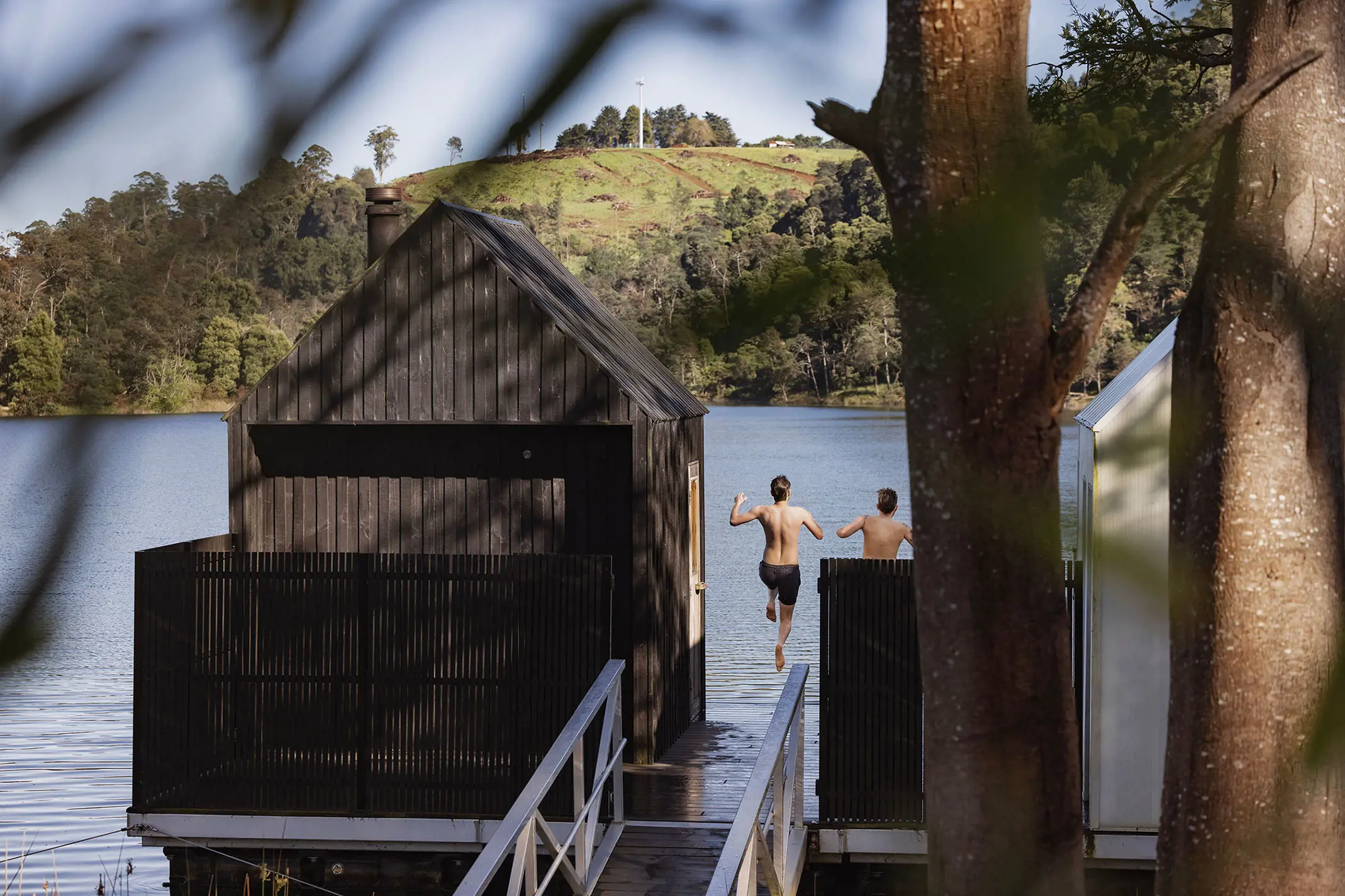 Two people jump into the cold waters of a lake from the decking in front of a sauna.